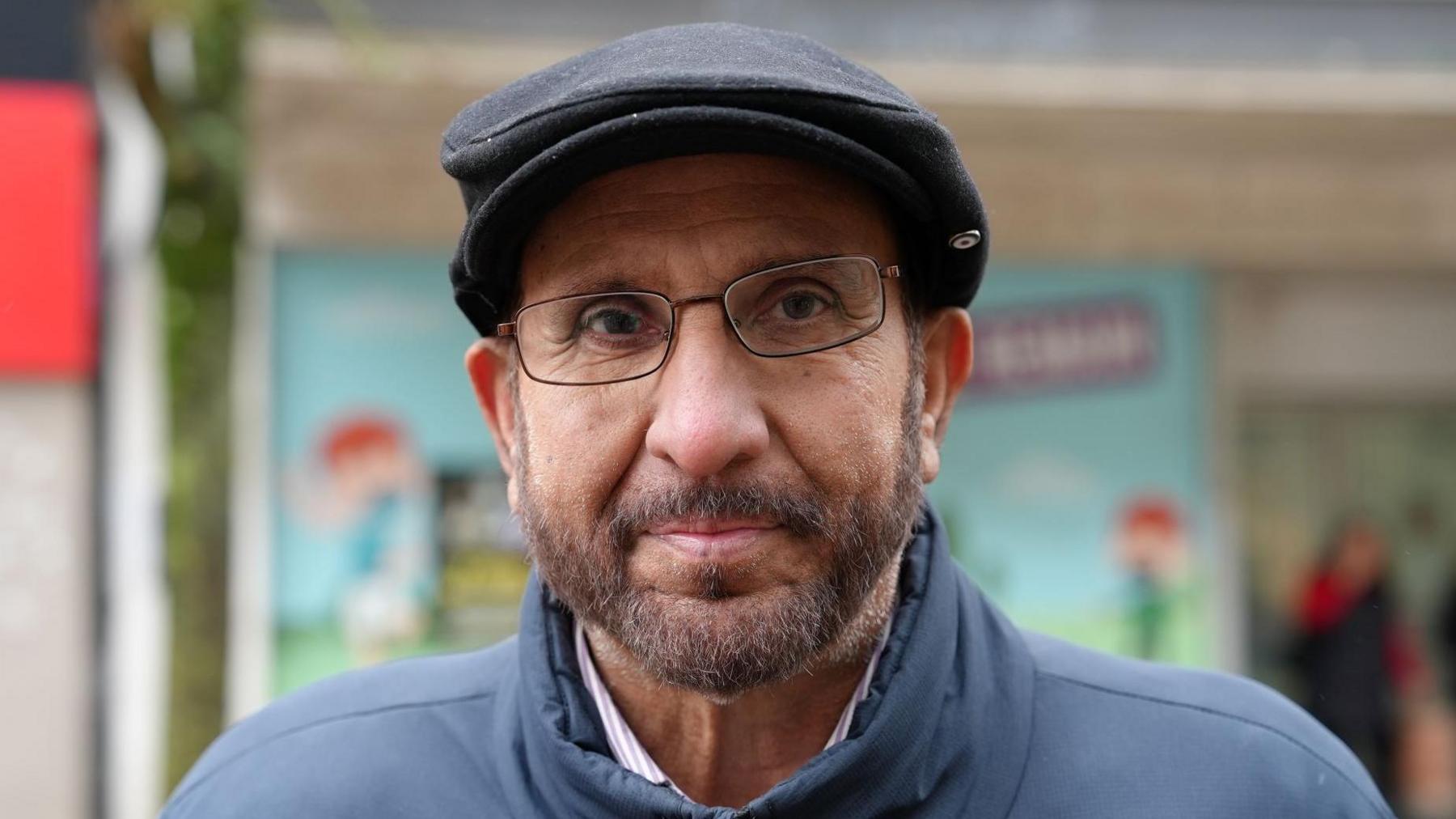 Khalil Rehman, who is wearing a black cap and glasses. He is offering a slight smile while wearing a blue coat, and standing on a street with a shop behind him.