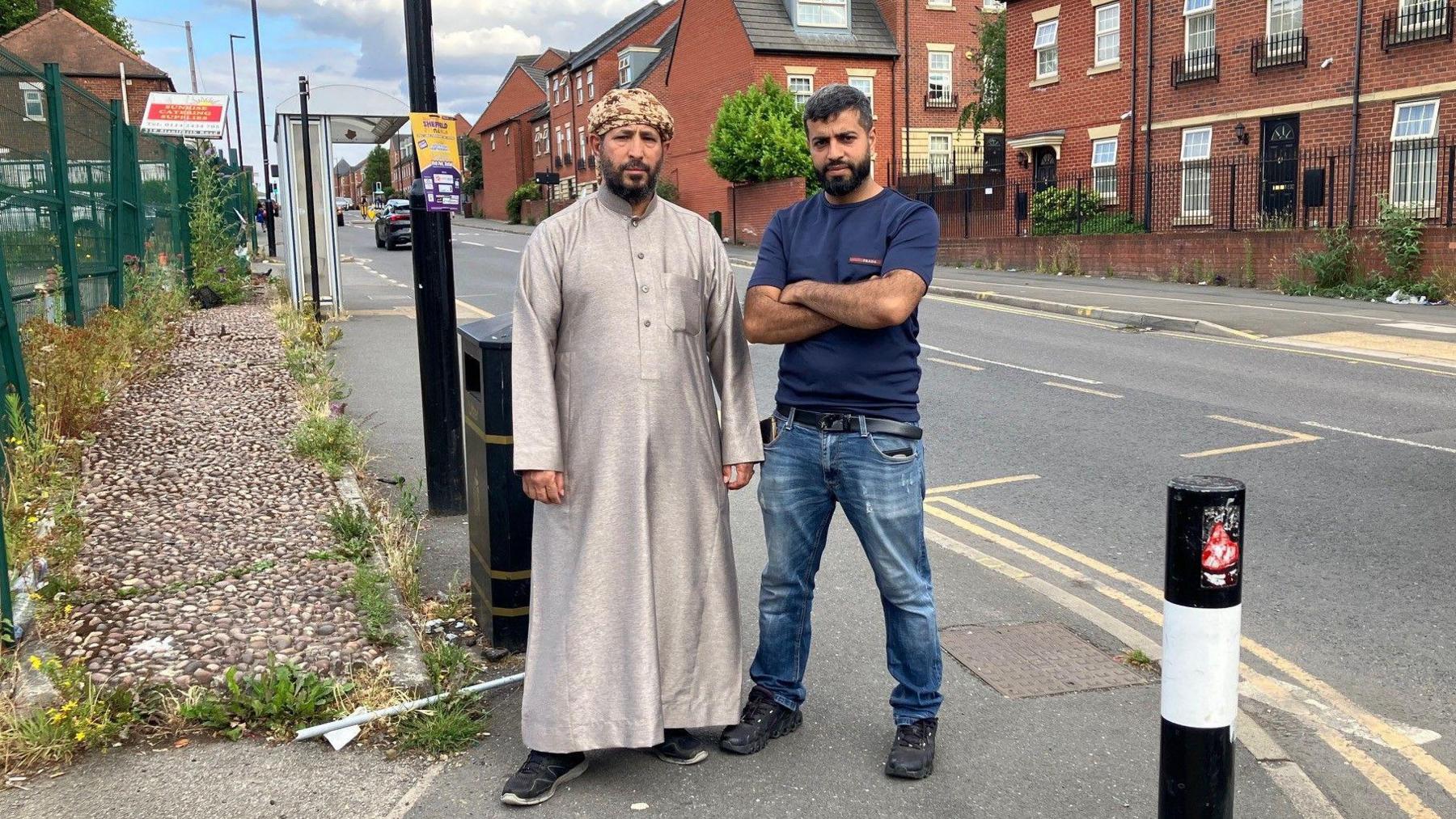 Two men stand on a street corner. They are surrounded by street furniture such as a lamp-post, a bollard, a bin and a bus stop. Some flats are in the background on the other side of the street.