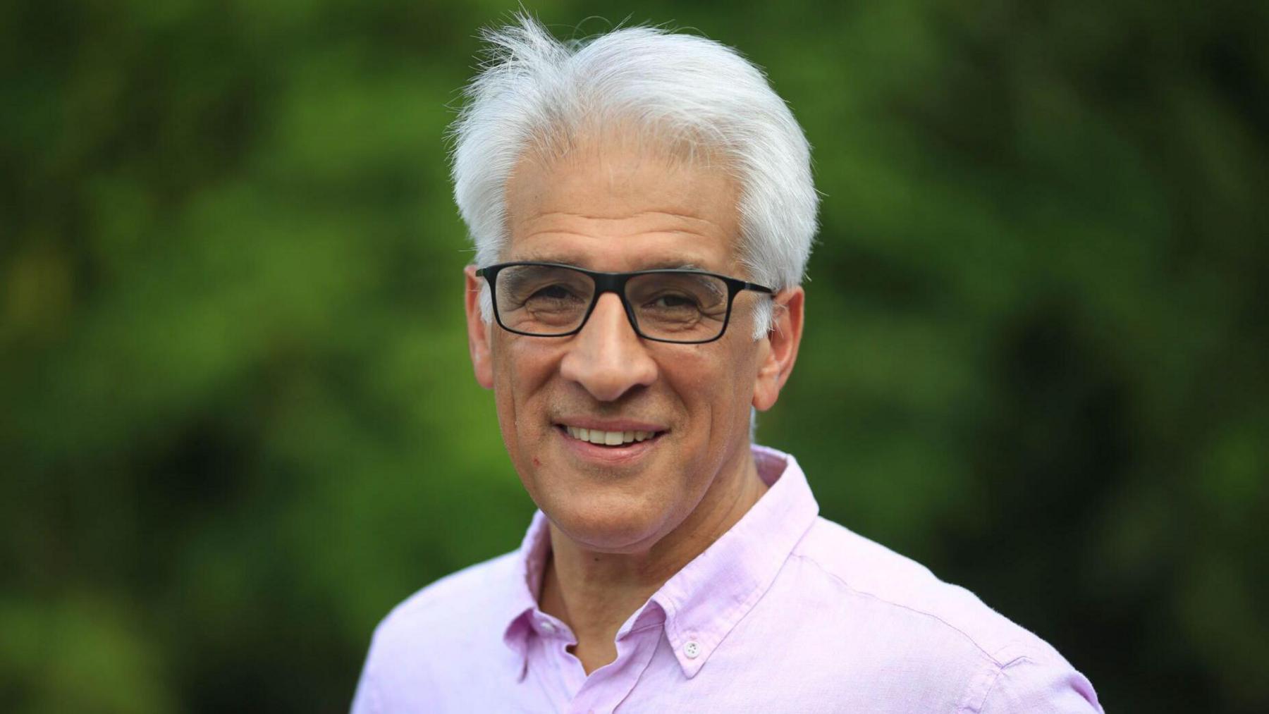 Mr Chalke with short, neatly styled white hair wearing a light pink button-up shirt. He is standing outdoors, with the background blurred.