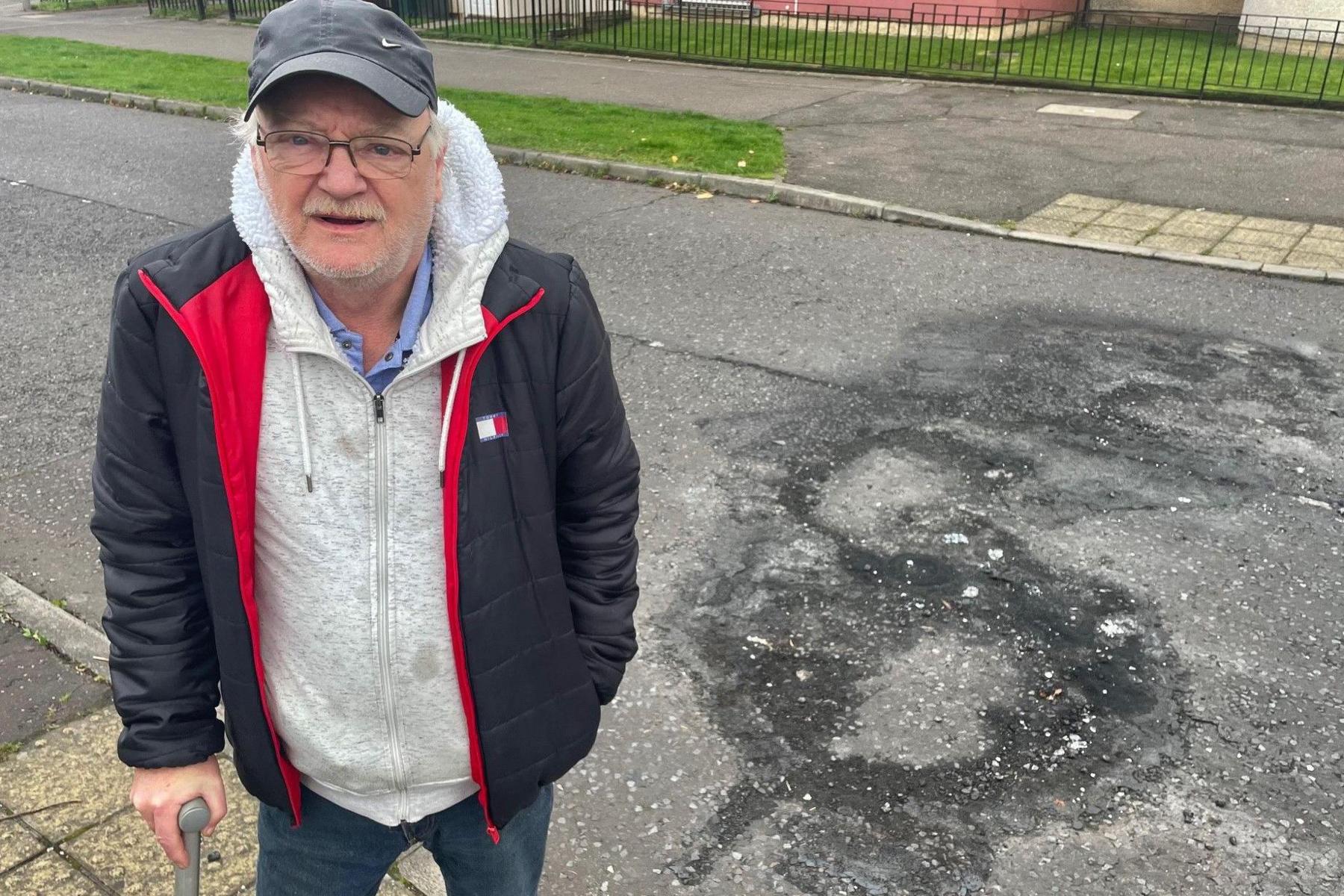 A man wearing a dark blue jacket and grey hoodie stands next to the road outside his home - there is a large burn mark on the road where a bonfire had previously been set