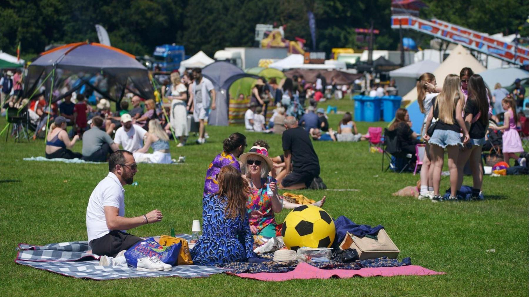 A man and two women sit on a rug enjoying a picnic at the Africa Oye event in Sefton Park in June, for which Sefton Park was also used. It is a sunny day and there are a number of other people sitting on blankets on the grass in the background.