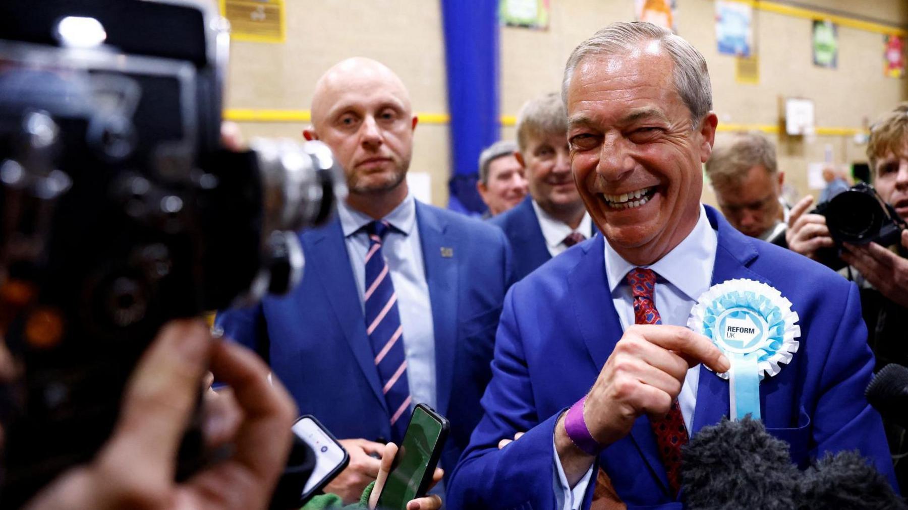 Nigel Farage on election night. He is in a sports hall wearing a navy suit with a blue Reform UK ribbon on.