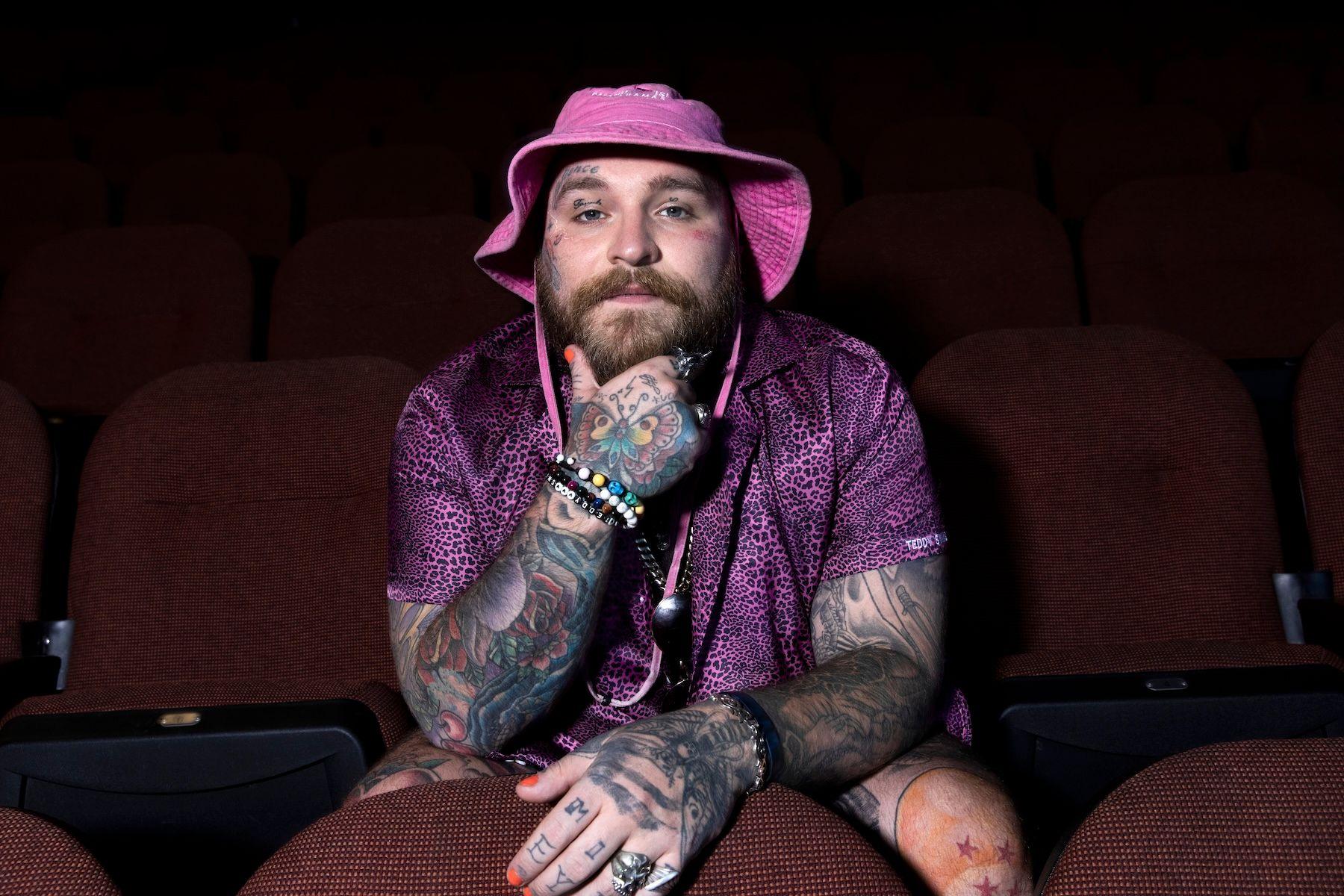 Teddy Swims rests his head in his hand, while sitting in cinema-style seats, wearing a pink bucket hat and a purple leopard-print style shirt.