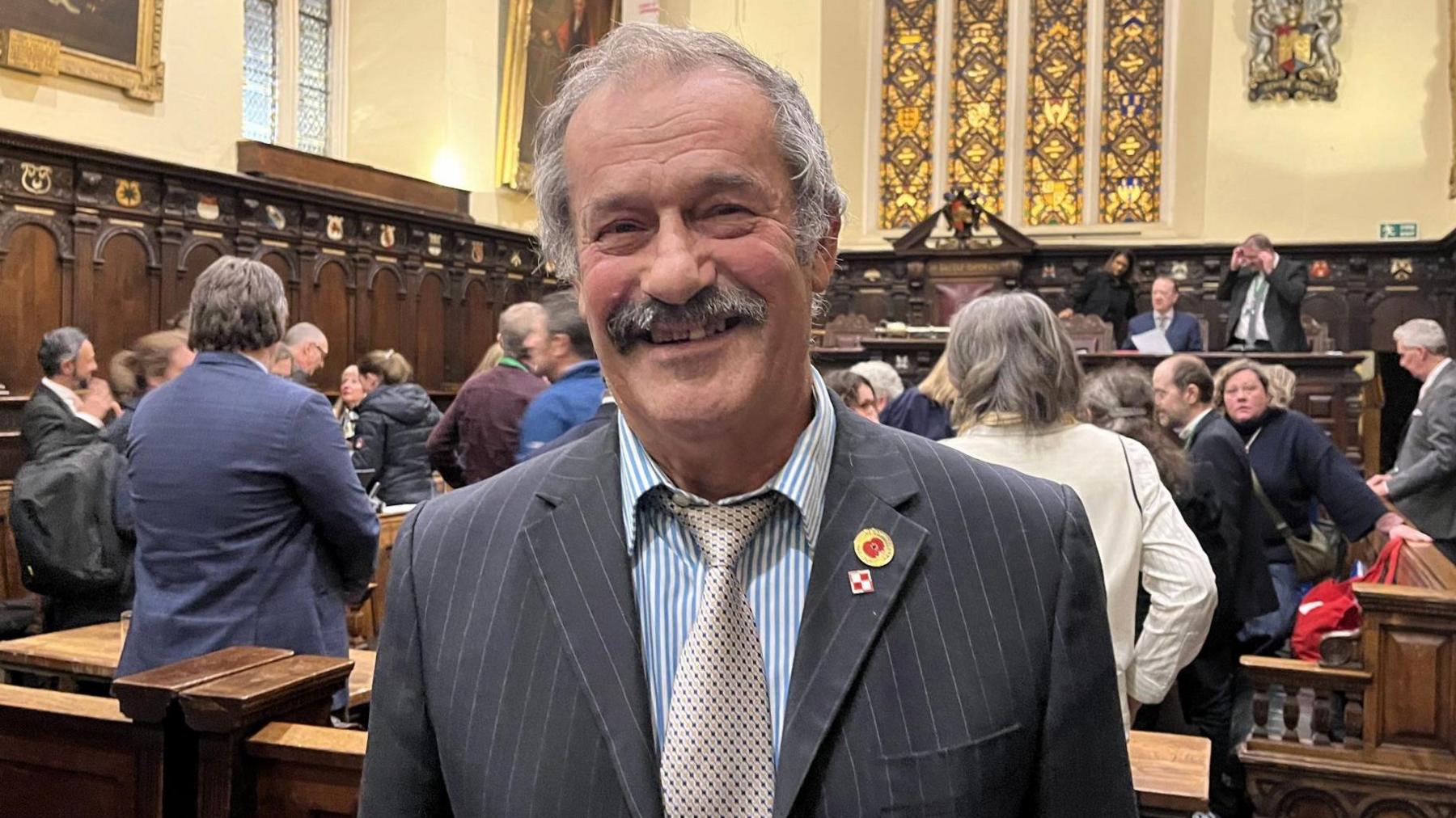 The image shows a man dressed in a suit and striped tie standing in a formal, historic-looking room. He has two pins on his lapel and appears to be attending an event or ceremony. The room is richly decorated with wooden panelling, stained glass windows, and framed portraits. In the background, other people are present and engaged in conversation.