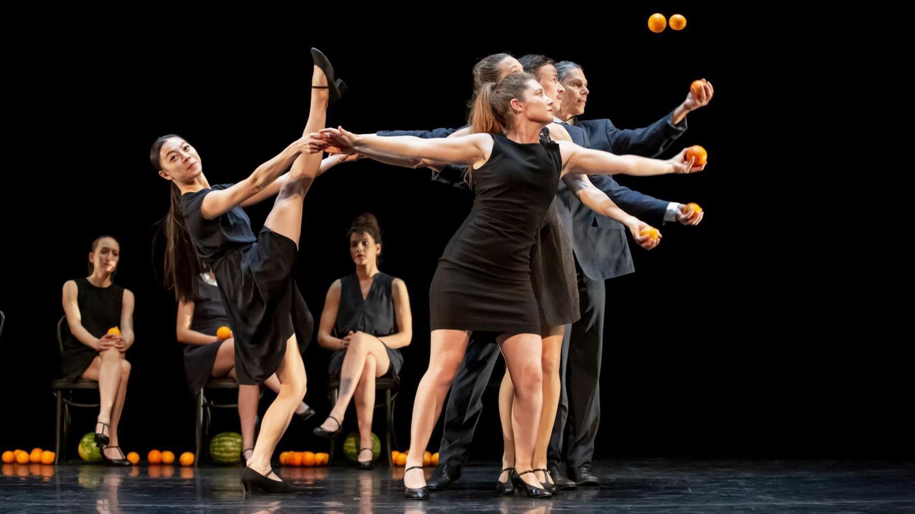 A group of men and women dressed in all black on a stage juggling oranges. One woman is performing a high kick while three other sit on chairs in the background.