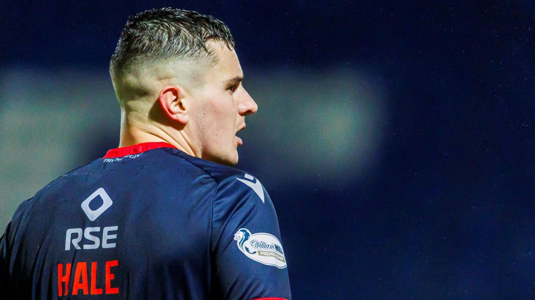 Ronan Hale is wearing Ross County's dark blue home kit with red flashes. On the back of his short is the name of the team's sponsor, RSE, in white letters and below that the player's surname in red capital letters. Hale is looking to his right.
