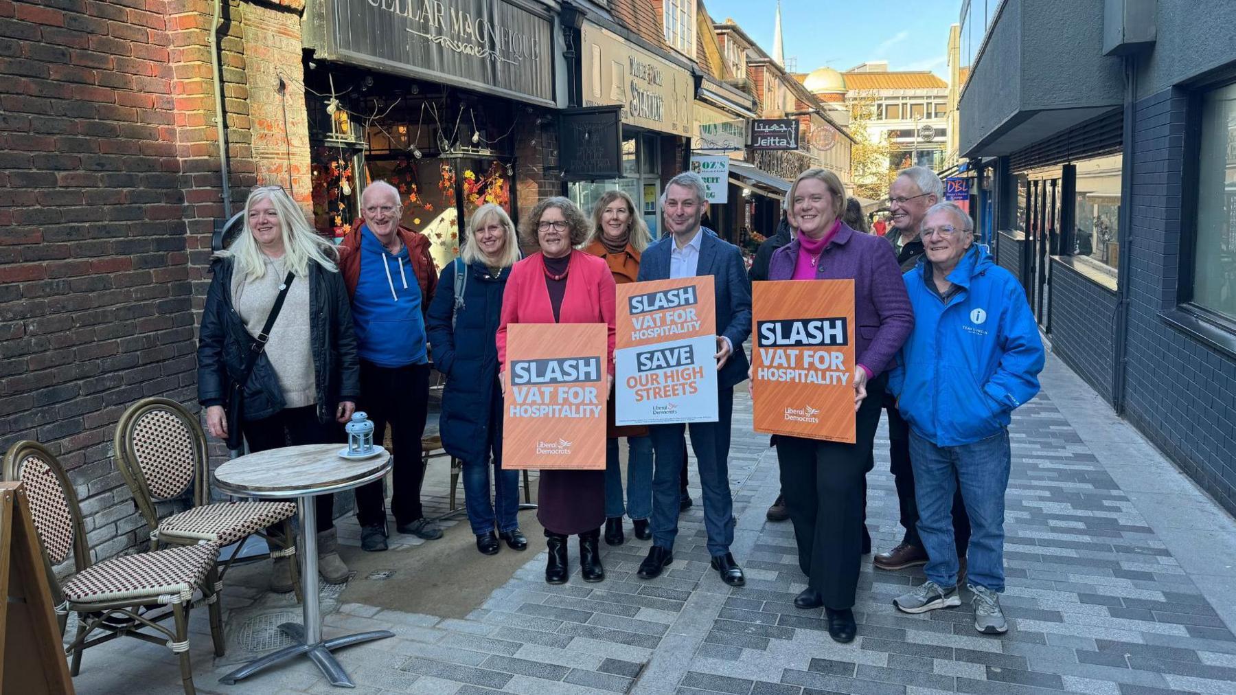 Woking town centre. In the middle of the picture is the leader of Woking Borough Council, Ann-Marie Barker, the MP for Woking, Will Forster and the MP for Guildford, Zöe Franklin. They are all holding orange signs calling for VAT to be slashed. Liberal Democrat campaigners and councillors stand behind them.