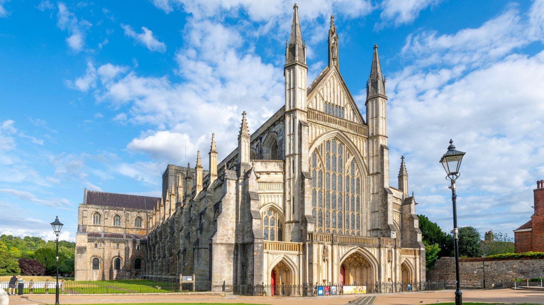 Winchester Cathedral, a large old building which has dozens of stained glass windows. There are lamp posts in the foreground, and areas of green space surrounding it.