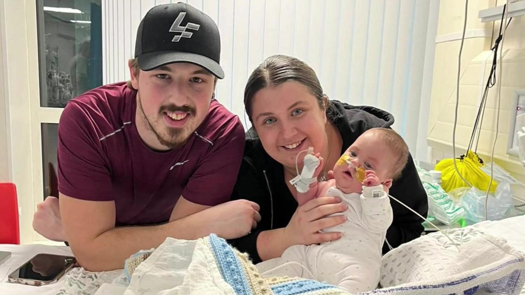 Jay and Lilly holding their baby Bobby, who has a tube in his nose. He is sitting on a blanket in a hospital bed.