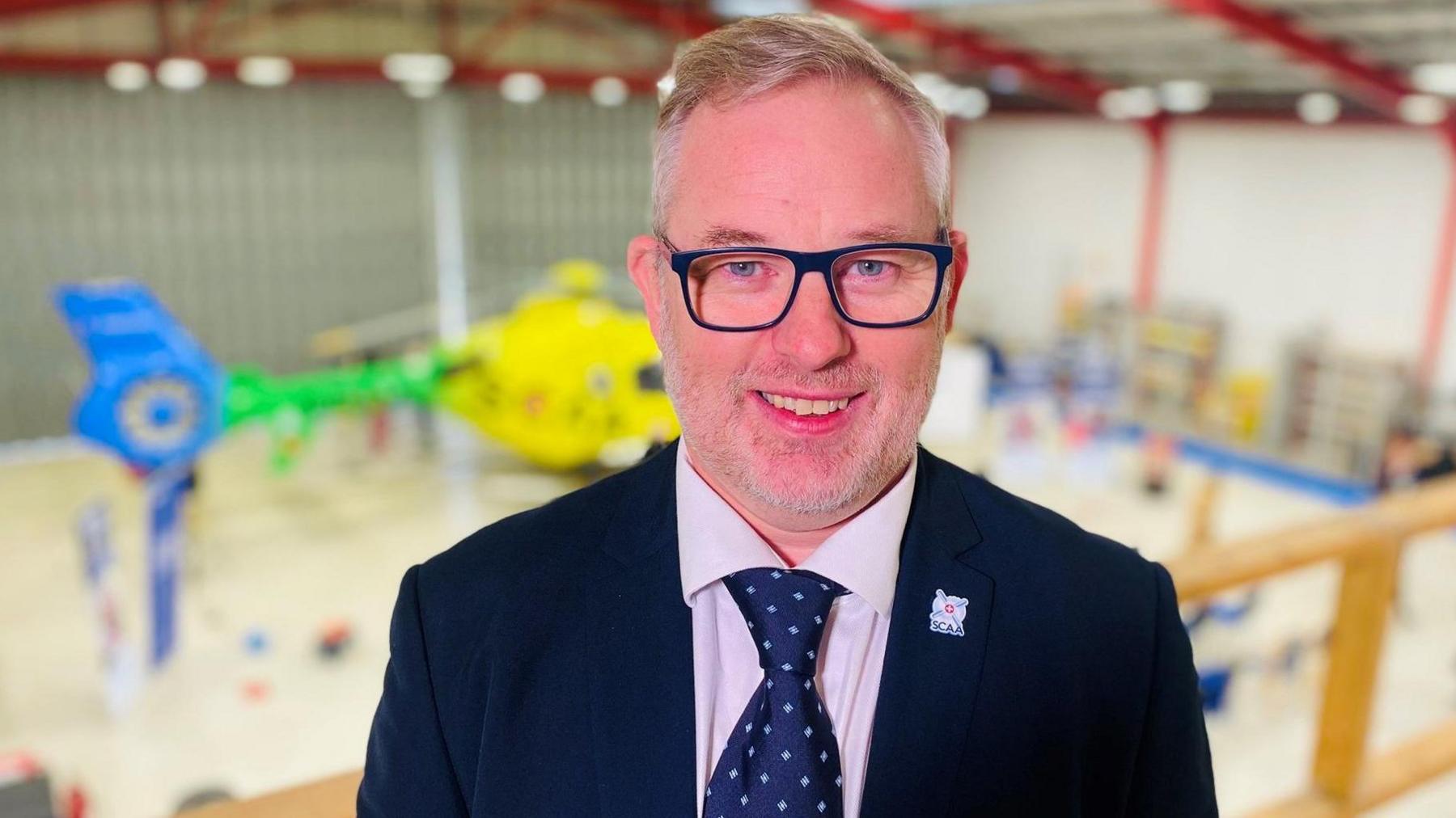 Man in suit, shirt and tie and glasses, with greying hair, smiling at camera, with a yellow, green and blue helicopter in the background in a hangar setting.