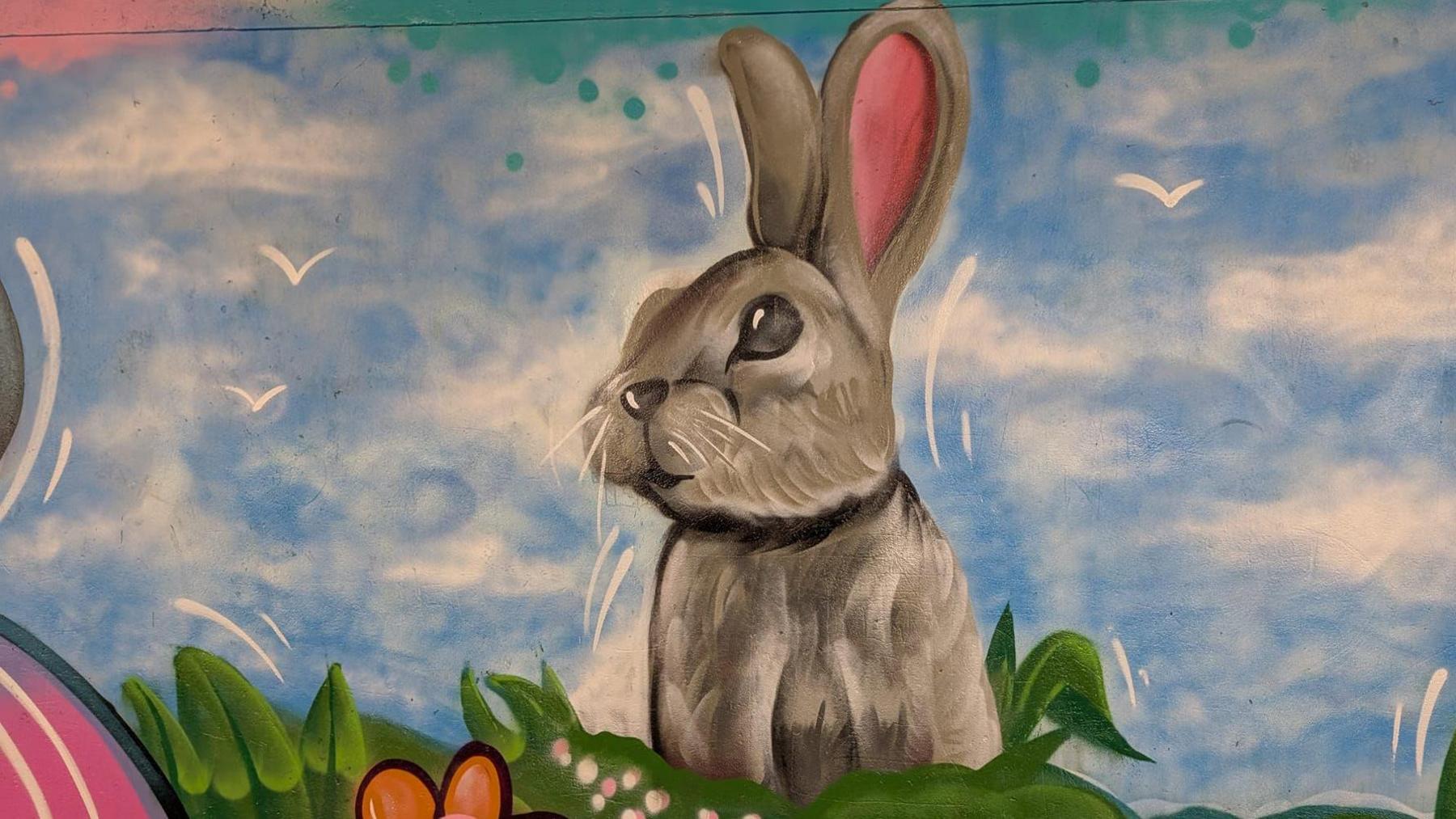 A grey painted rabbit on a blue sky background, with green grass and orange flowers underneath