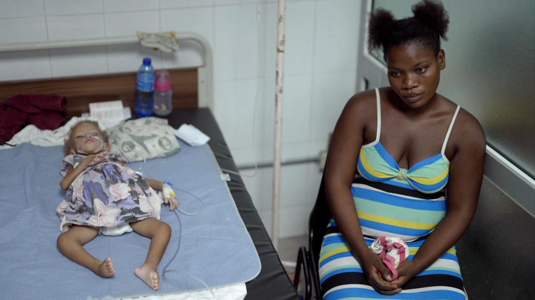 A small emaciated child lies in a bed hooked up to a drip while her mother sits by her bedside. The mother is clutching a cloth in her hands.