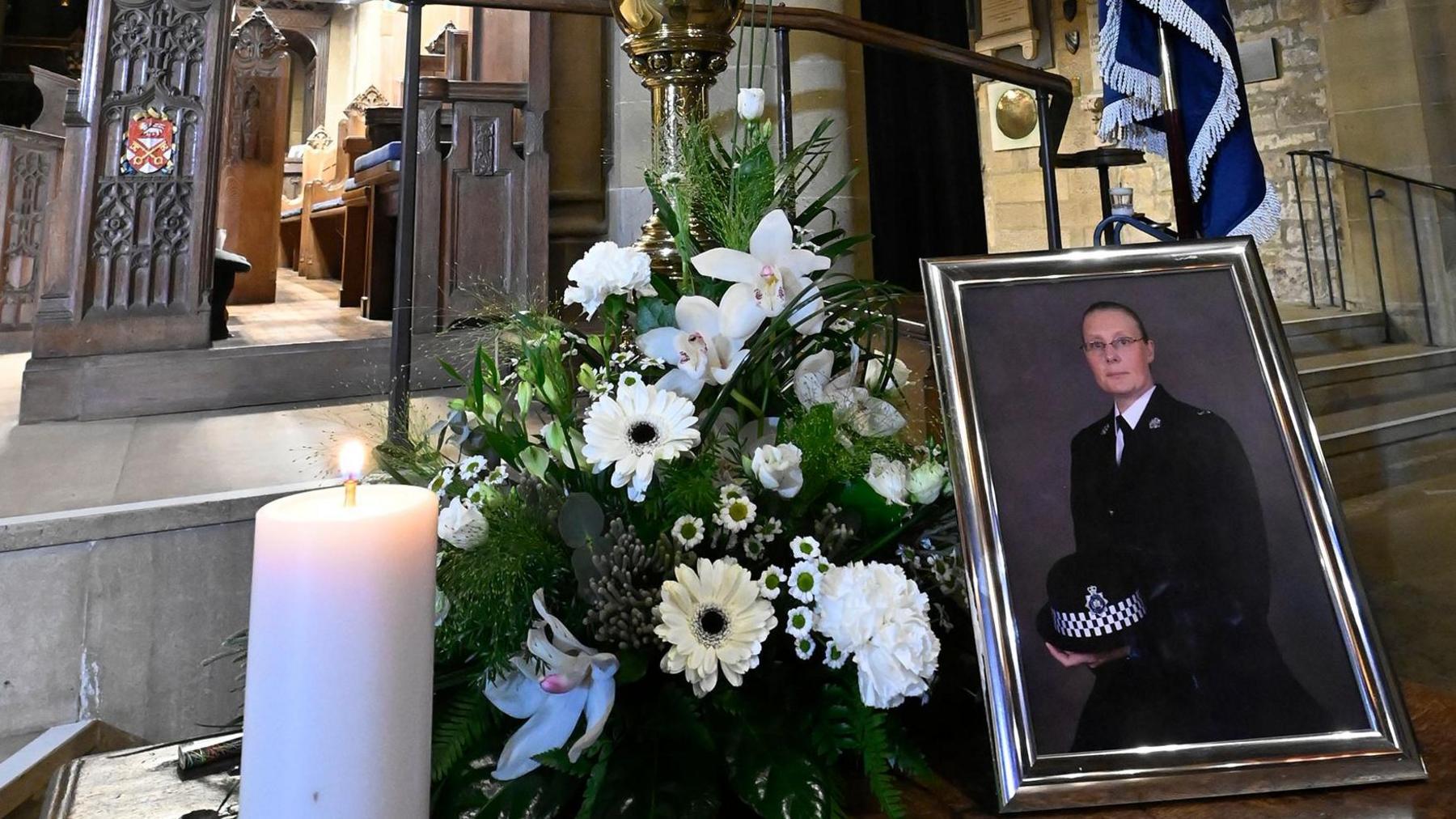 A framed photo of a police officer in uniform, next to flowers and a lit candle. The items are in the front of a cathedral.