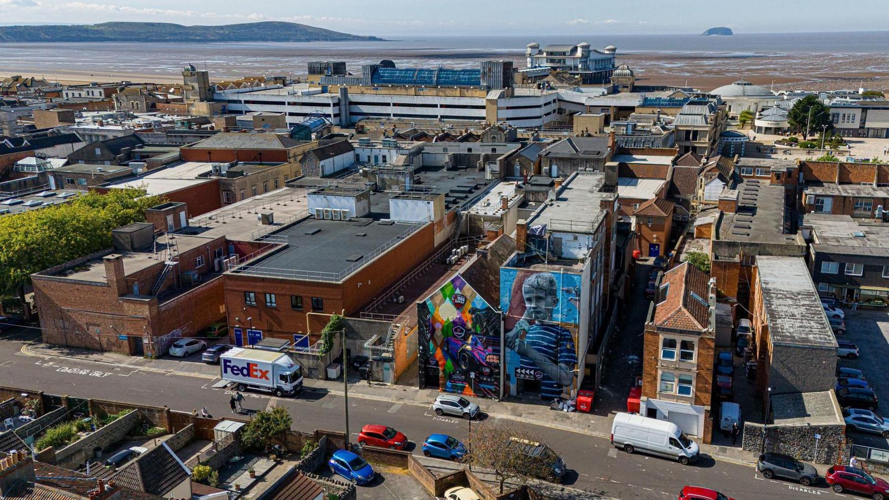 A large street mural in Weston-super-Mare is seen from a drone shot, with the town's pier and coastline visible in the distance. The mural is part of the Weston Wallz festival