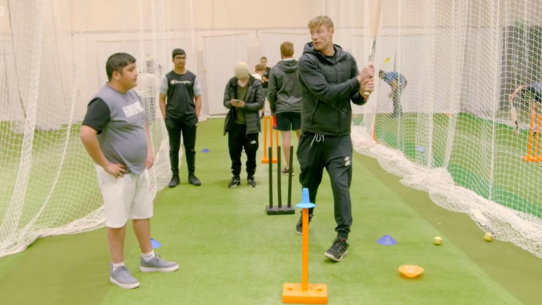 Freddie Flintoff is wearing a dark coloured tracksuit and is holding a cricket bat in cricket nets and is speaking to Umar Mahmood who is wearing white shorts and a grey top with grey pumps.