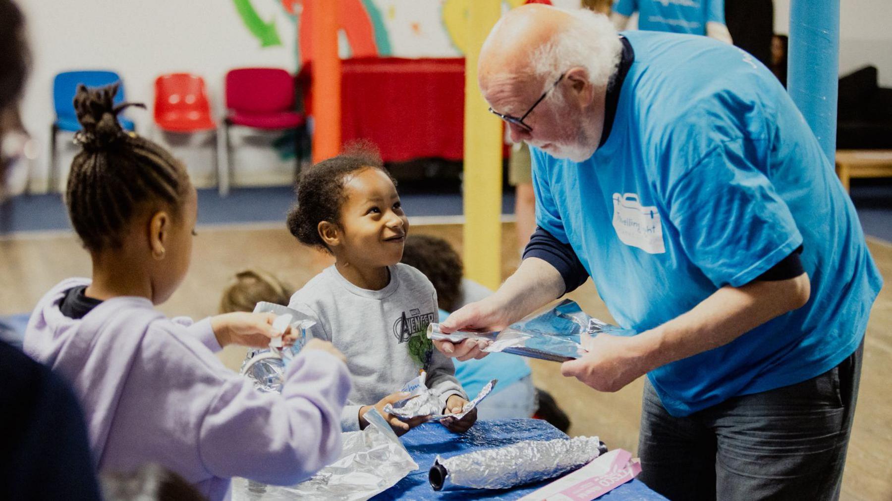 Two children making arts and crafts at a table with a man from Travelling Light. There is a roll of foil on the table. The children are making things out of it. One of them is looking up at the man and smiling. 