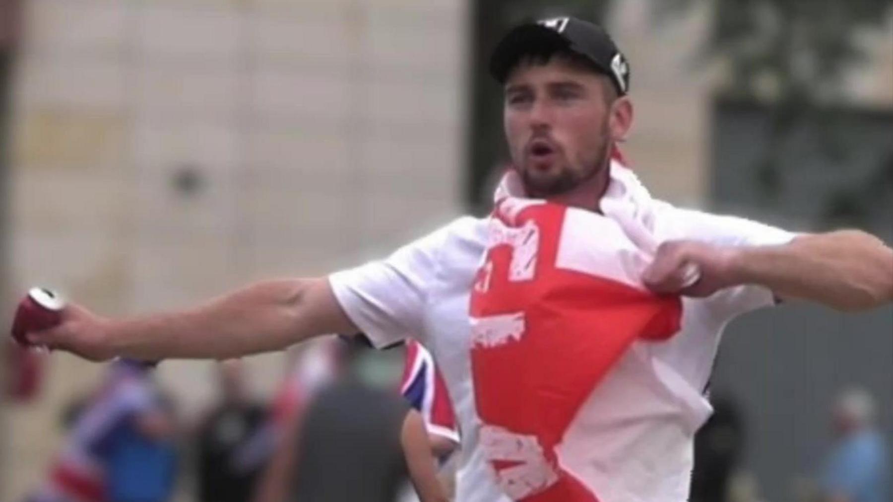 Still of Kirtley during the riot. He is wearing a white tshirt and black baseball cap, with a red and white England flag draped round his neck. He is a young man with a black beard, and is holding his arms as if he is chanting