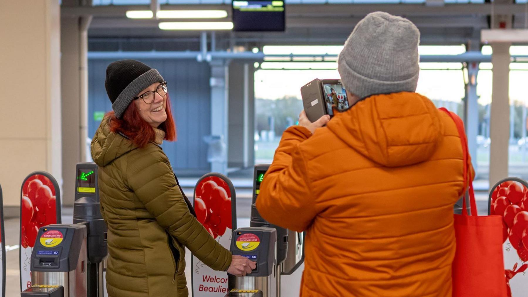 A woman wearing a coat and a hat smiles as she uses her ticket to get through the barriers. She is being photographed by a person wearing an orange coat.