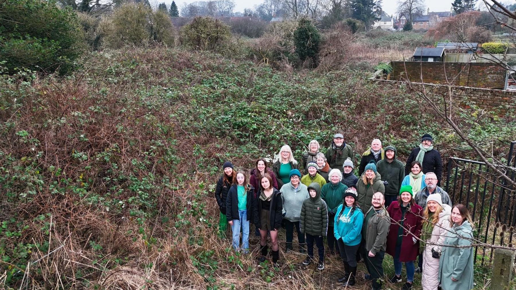 A photo taken at an angle looking down on the of the Friends of Driffield Sustainable Park - a campaign group of about two-dozen men and women standing on an overgrown patch of land in Driffield. They are dressed in winter clothing, surrounded by tall bushes and greenery, with garden fences to the right.