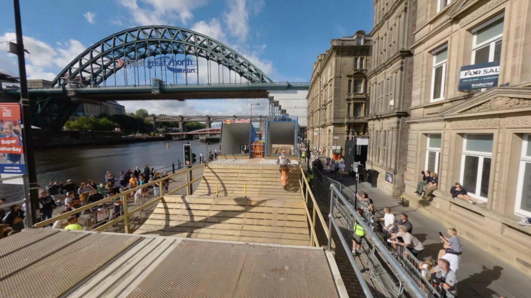 Drone shot of a the back view of a motorcyclist riding down on a specially built course on Newcastle's quayside, consisting of a series of wooden ramps. People can be seen cheering on either side, with the Tyne Bridge spanning the river in the background.