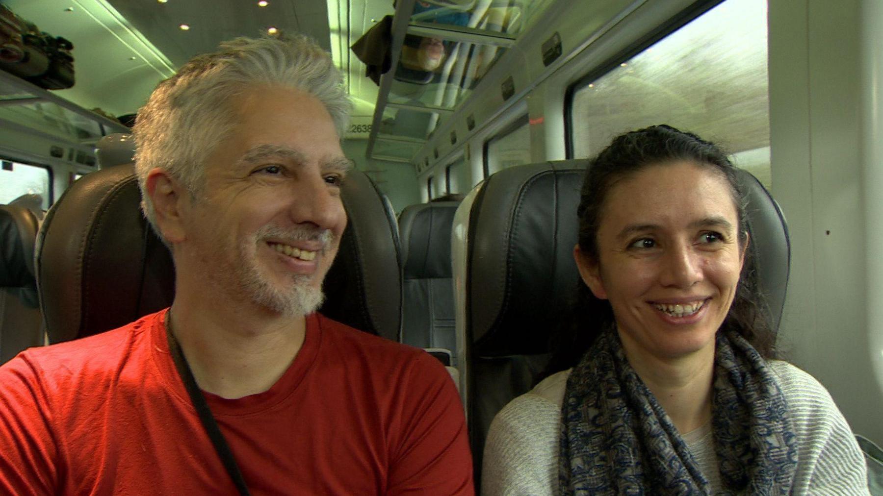 Ural (male) and Ayse (female) sitting on the train. Ural has white hair and beard and wearing a red t-shirt. Ayse has dark hair tied back and wearing a grey jumper and blue/grey scarf. Both are smiling.