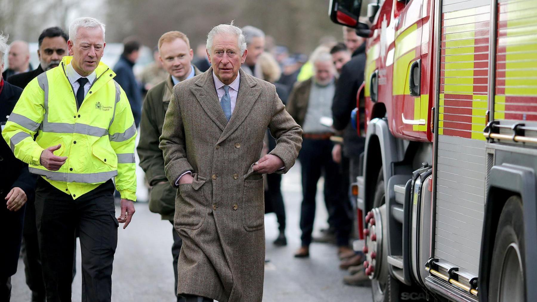 A smartly dressed man in a beige winter coat walks towards the camera in discussion with a man in a bright yellow jacket. They walk alongside a fire engine.