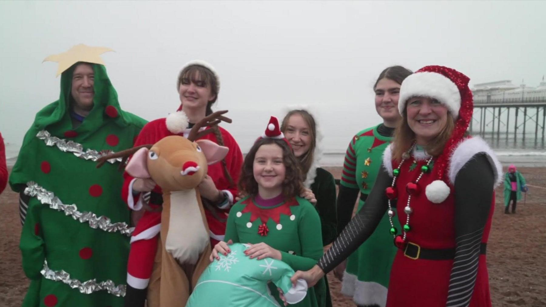 A family seen in Christmas fancy dress stood at Paignton Seafront. There are six people in the image. One person is dressed as a Christmas tree, a second as Santa Claus while the others are dressed as Christmas elves while a sixth is seen riding a fancy dress reindeer. 