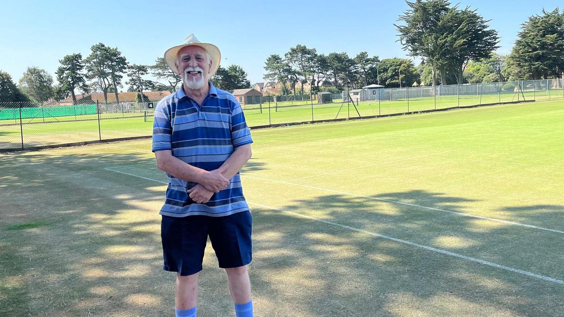 Organiser Chris Holt stands in front of a link fence inside of which are lots of tennis courts marked out with white lines and mesh nets. Chris wears a blue horizontally striped t-shirt, dark blue shorts and blue socks. It's a very sunny day so he has a sun hat on to shade his face. He has white hair and beard. 