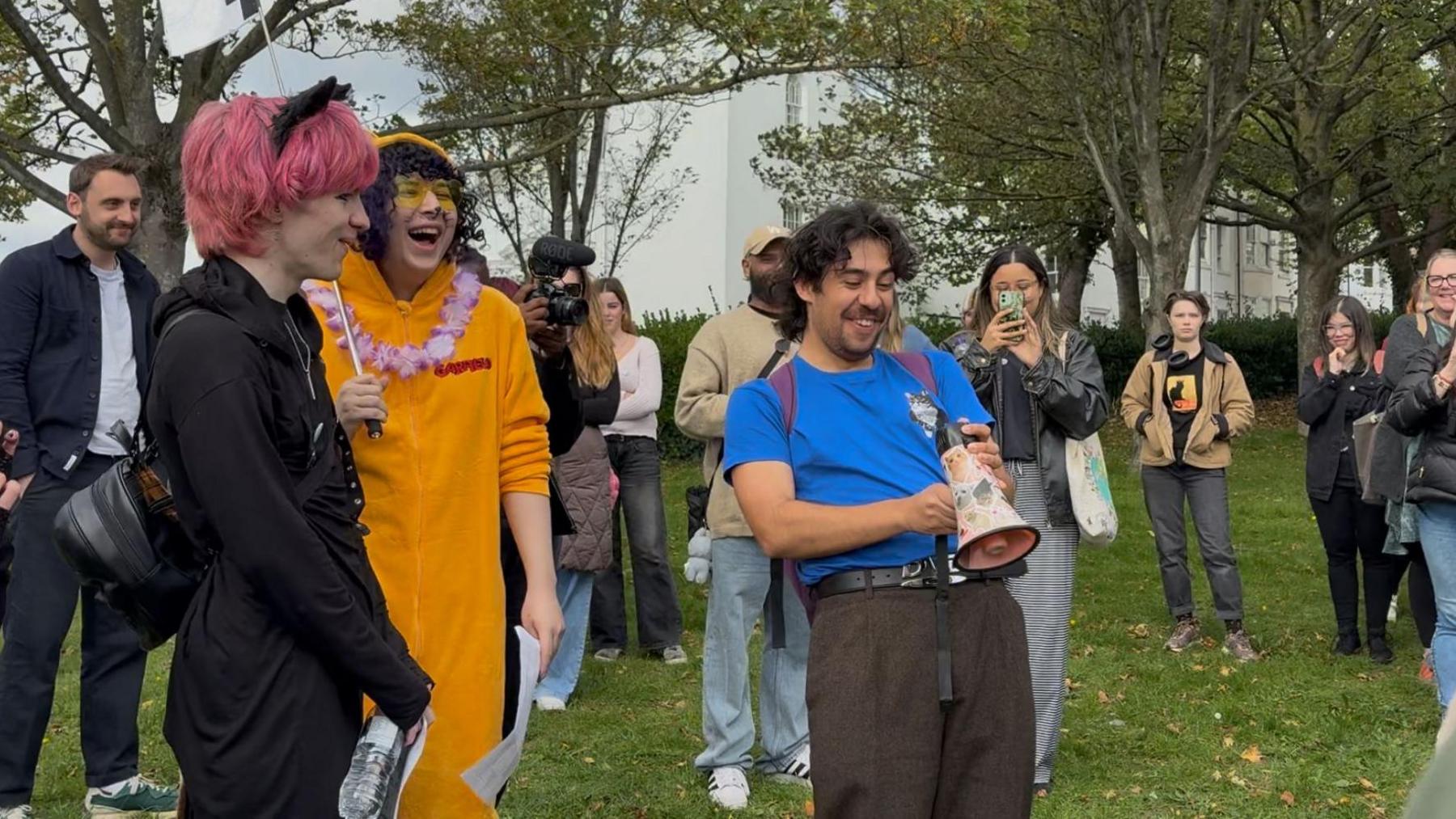 Two volunteers (left and middle) and Brighton Cat Tour organiser, Stefan Gutierrez (right), laugh in the middle of the crowd while having technical issues with their megaphone. The two volunteers are dressed in cat-themed costumes, one with dyed pink her and black cat ears, the other with a cat onesie, painted whiskers and floral necklace.