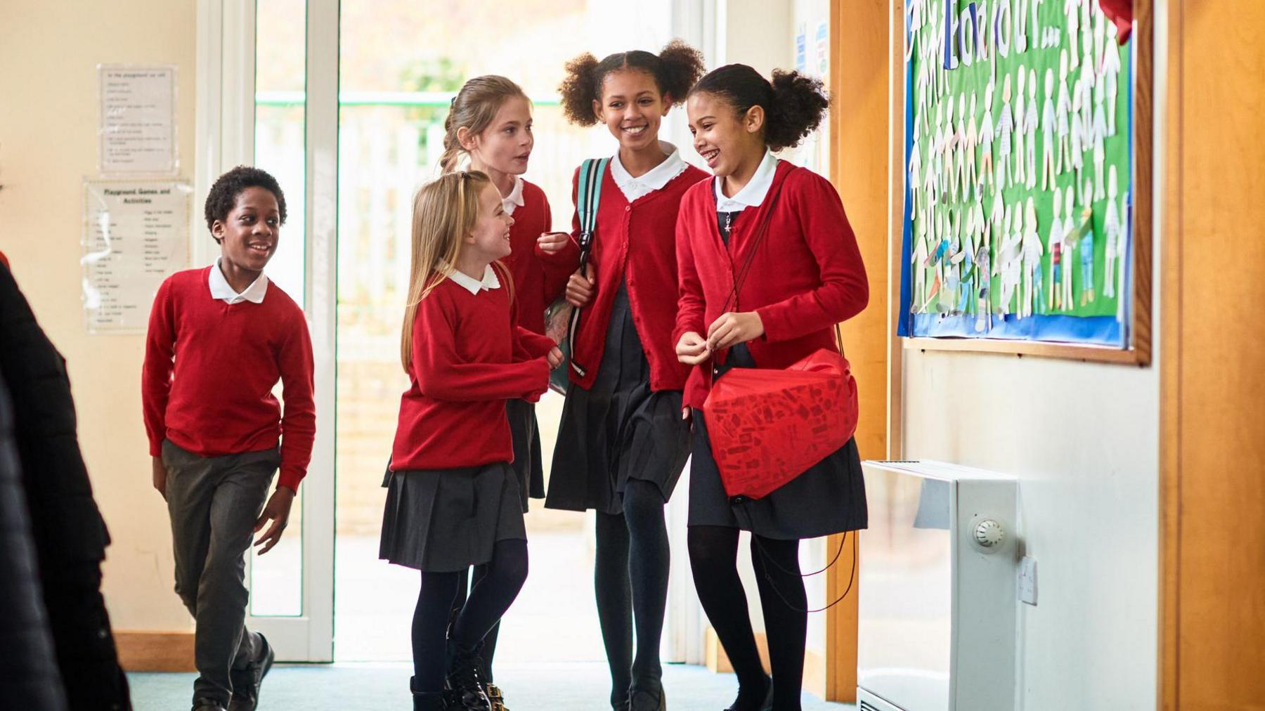 A group of young school girls talking in the corridor, with a young boy walking past. The girls are all wearing their school uniform which is a red cardigan, white shirt and black skirts with black tights. The boy is wearing the same cardigan and shirt but with black trousers.