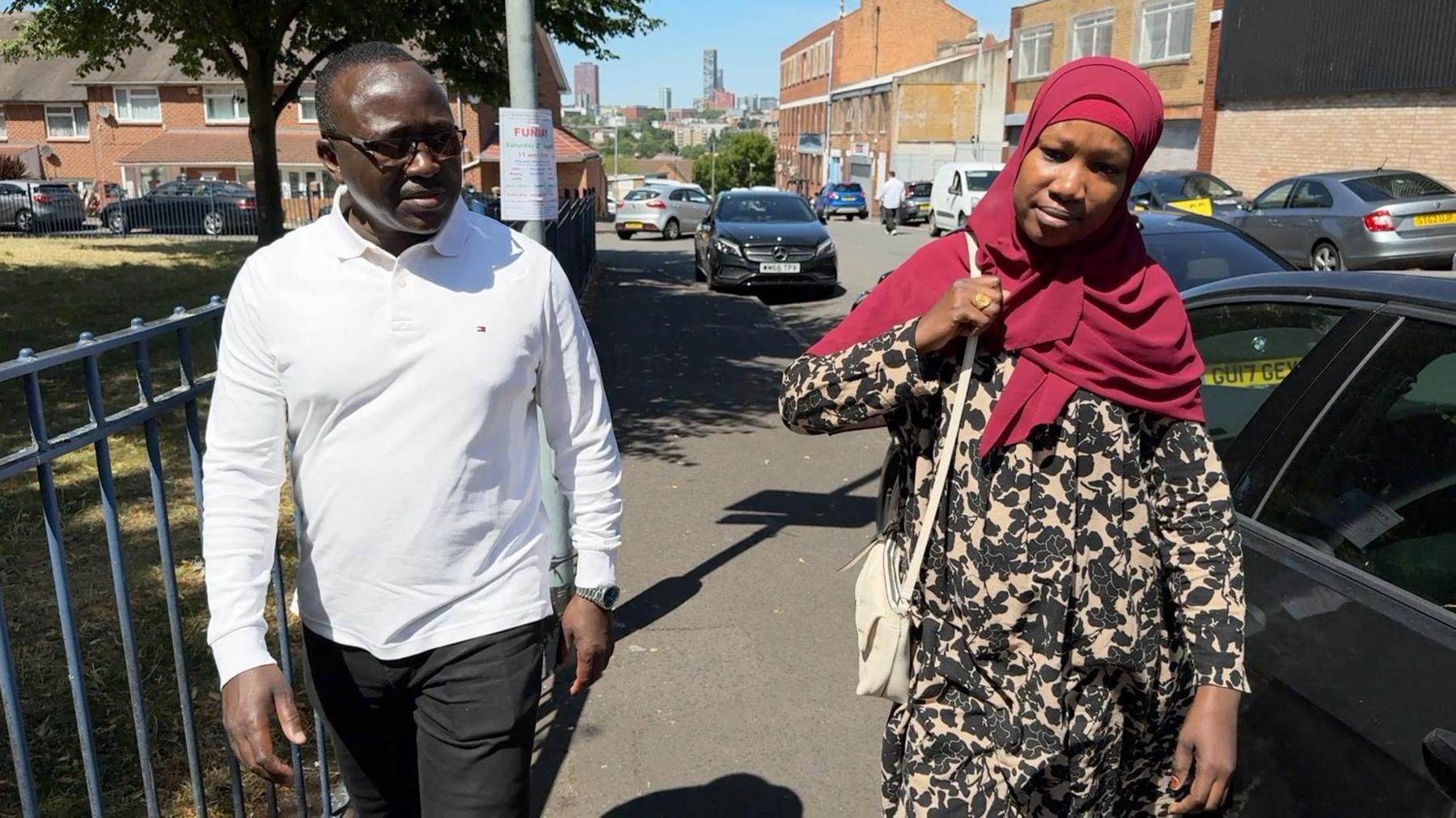 Babiker Yahia wearing a white long-sleeved top and black trousers walks down a pavement next to his wife, Sara Salih whose head is covered with a red scarf. She is wearing a brown coat with black pattern and a white handbag