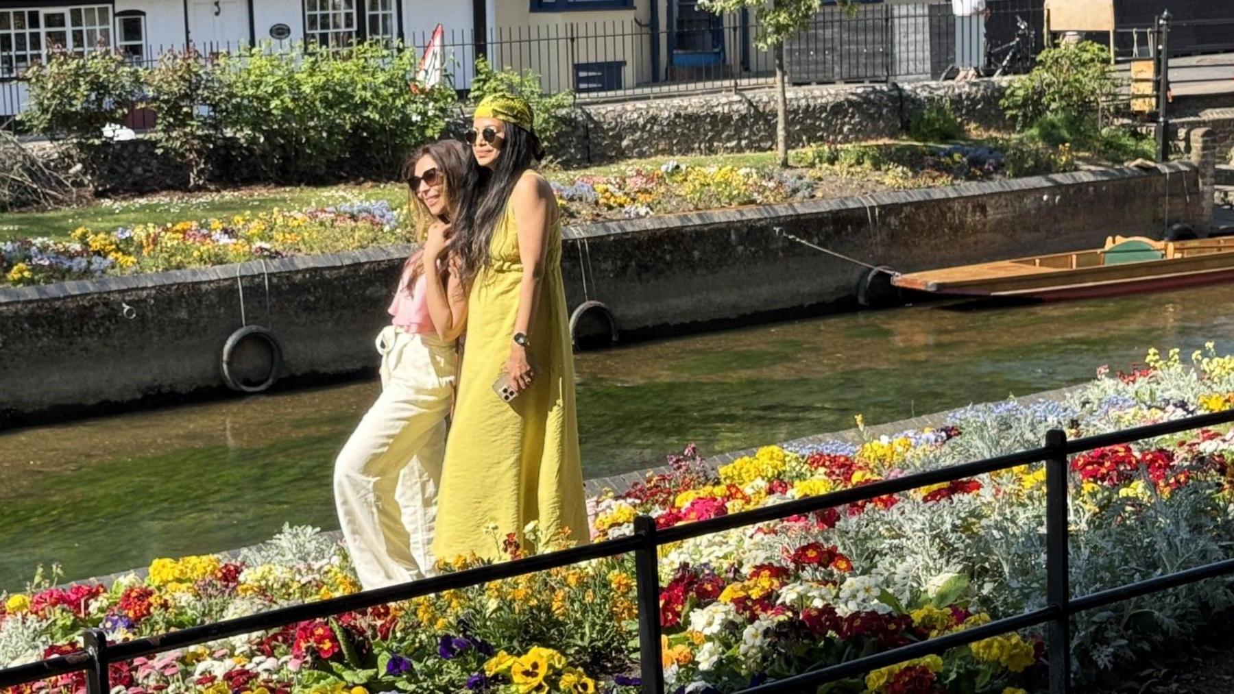 Two women, one wearing a cream top and trouser set, the other a yellow sun dress, pose for a selfie in the bright flower beds of Westgate Gardens, just in front of the River Stour, with a moored punt behind them.