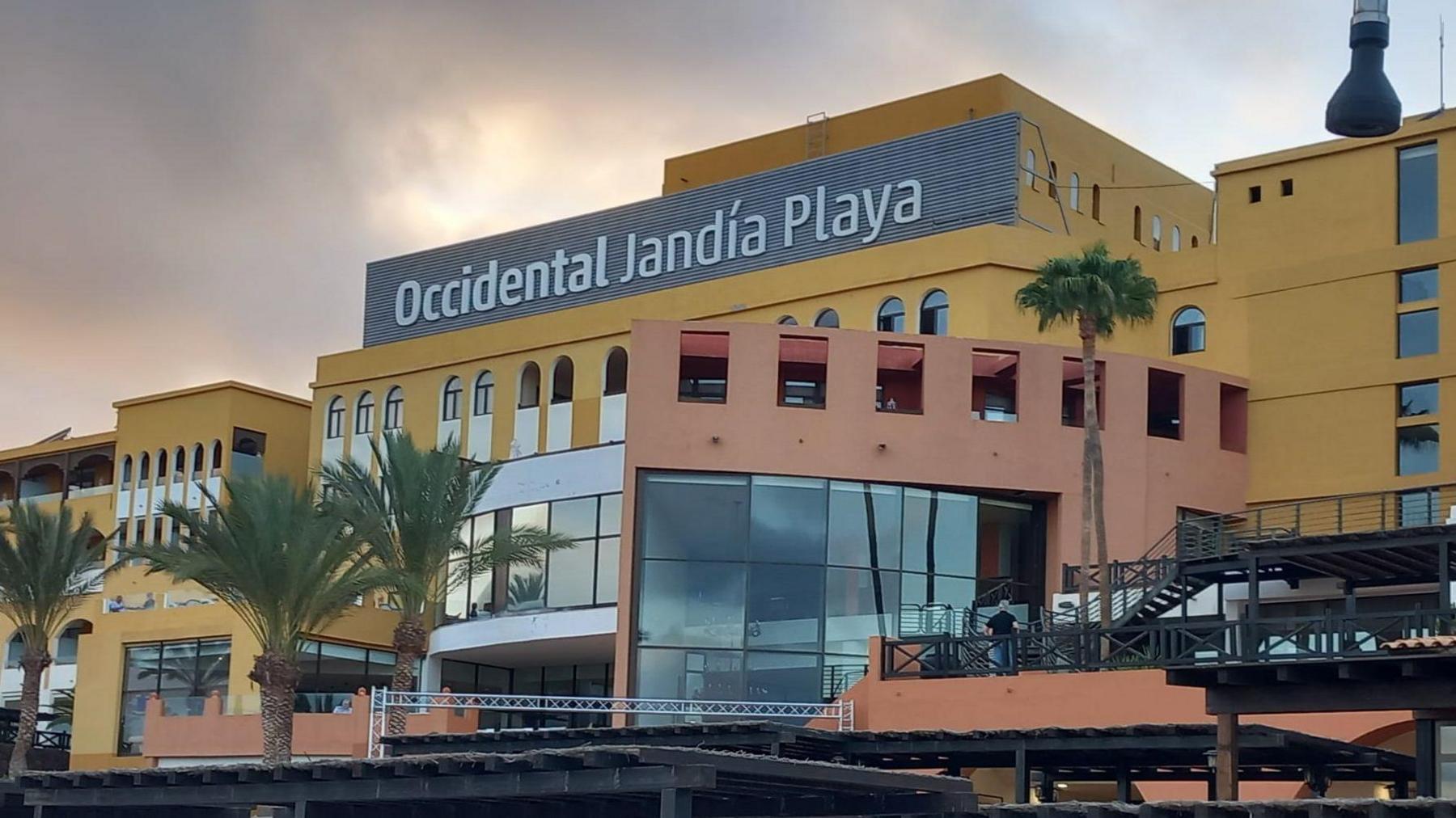 A general view of the front of a palm-tree lined hotel building, with a yellow fascia and a large sign with 'Occidental Jandia Playa' spelled out in white lettering.