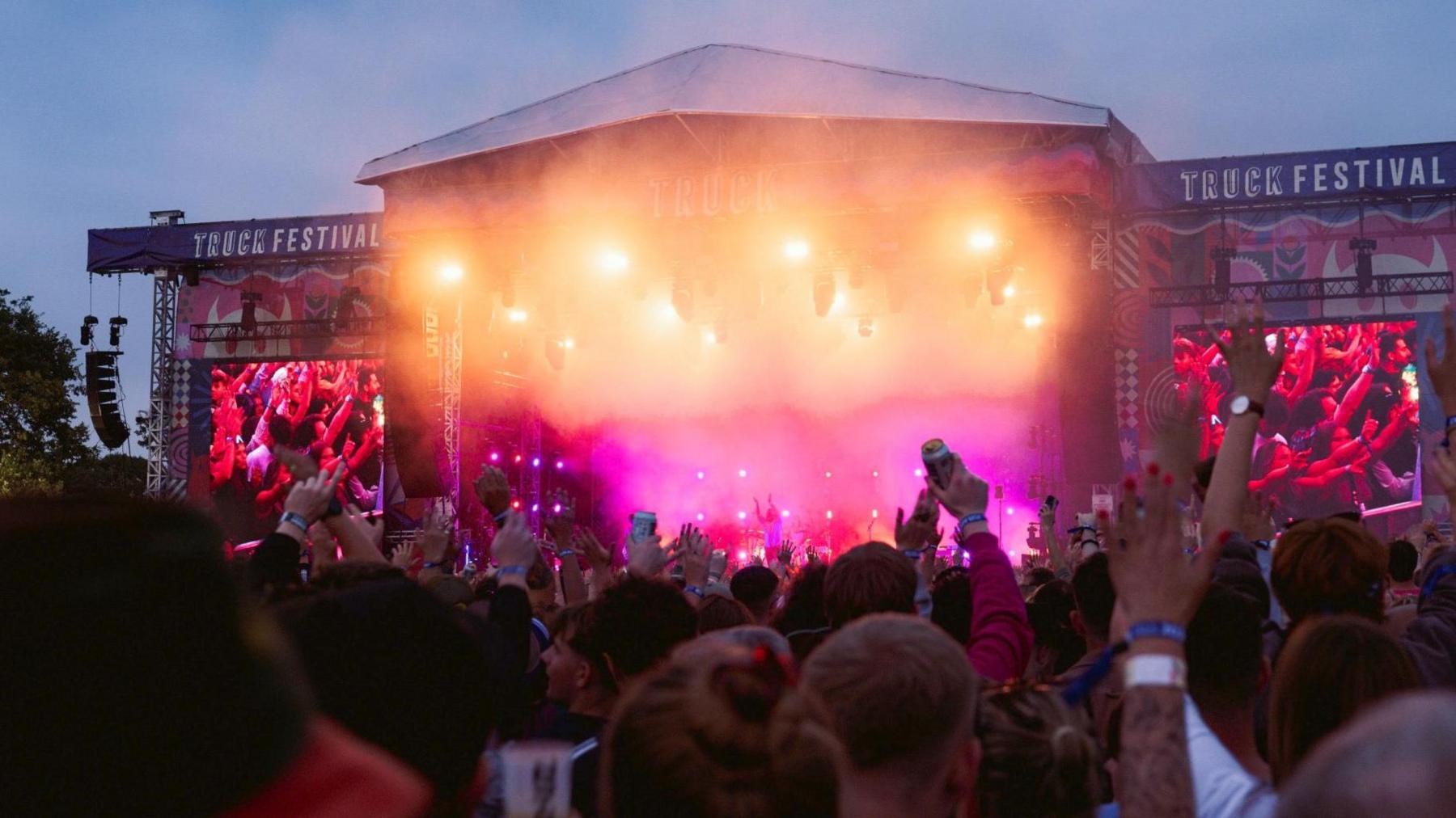 A view of the Truck Festival main stage from the crowd. People's hands are reaching up in the air and the stage is lit up in red and yellow bright lights.
