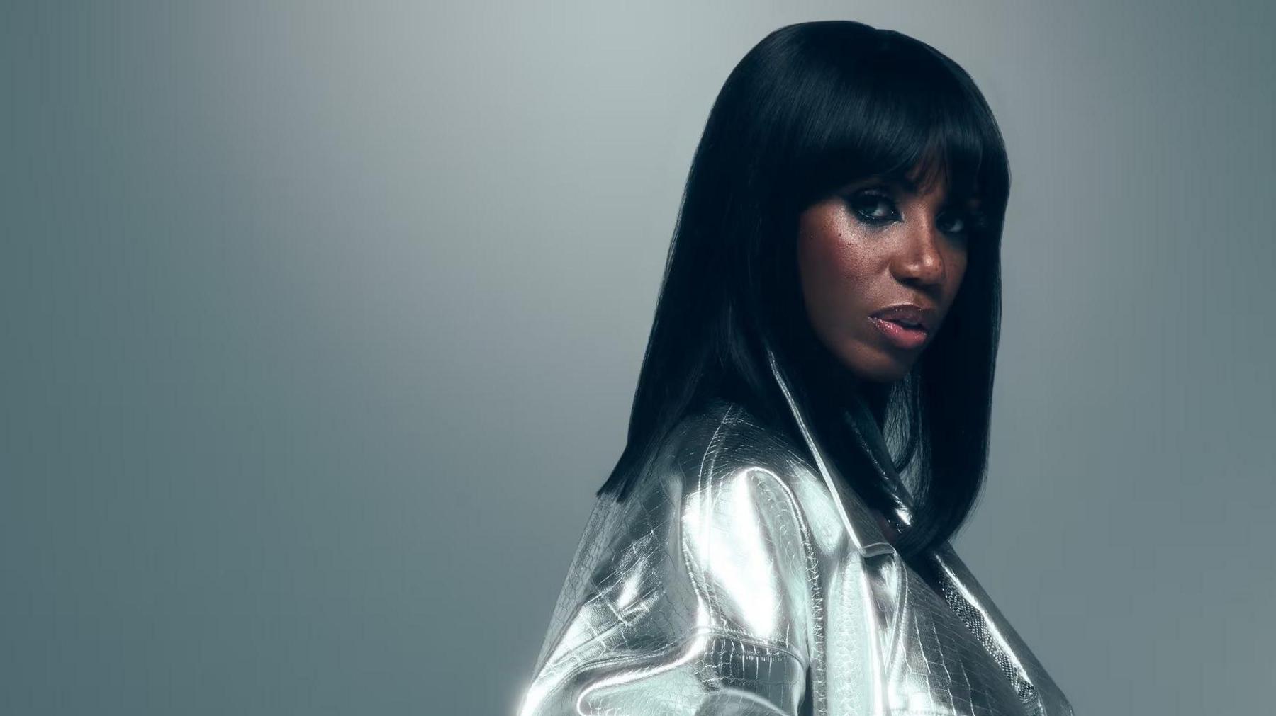 Promotional photo showing Shaznay Lewis against a grey background in a bright silver coat