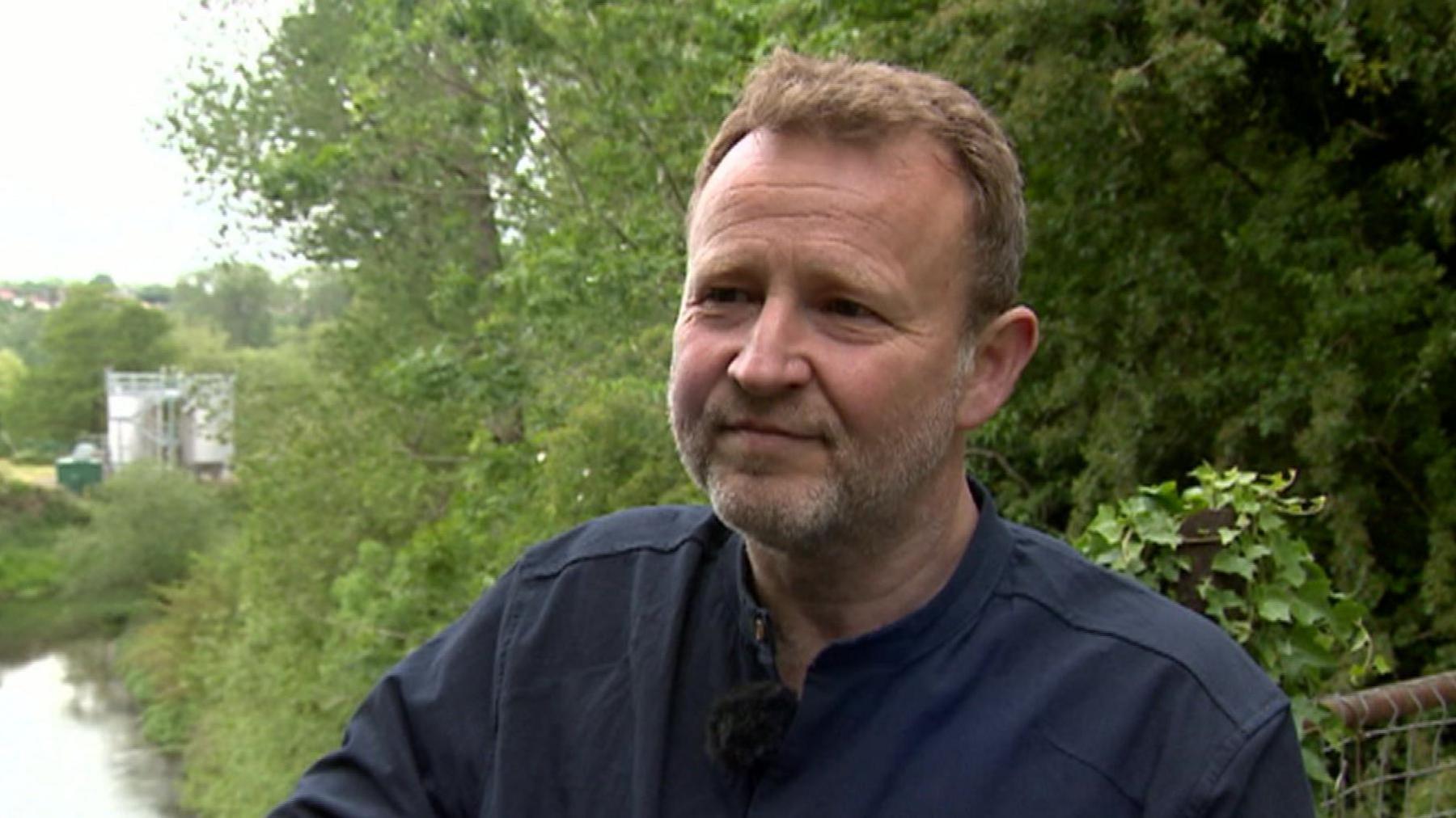 A man with short, light brown hair and beard and wearing a navy blue shirt stands on a bridge over a river. Foliage and a water treatment works is visible in the background.