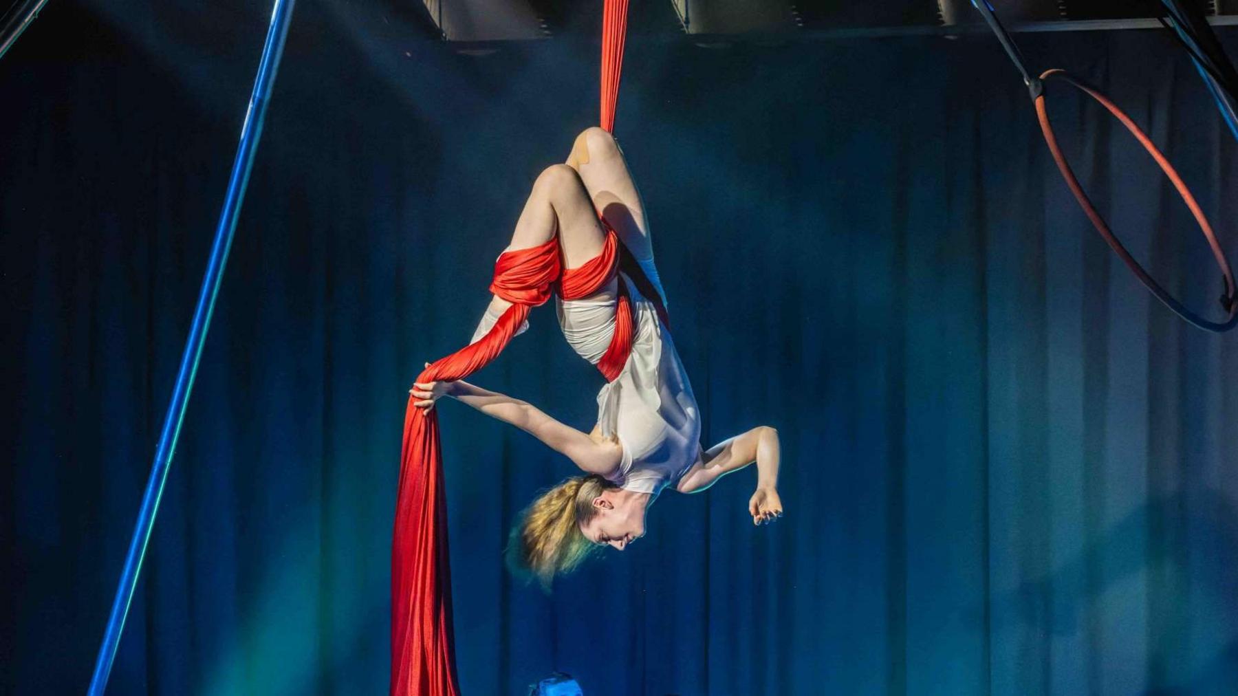 An aerial performer is on stage, hanging upside-down from a red piece of fabric. Her long blonde hair is in a ponytail.