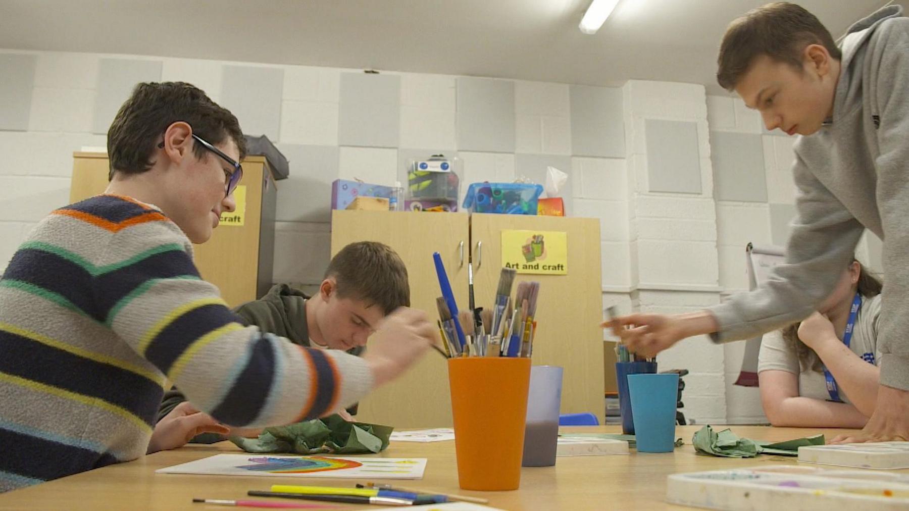 Teenagers round a table with paintbrushes, cups and paintings.