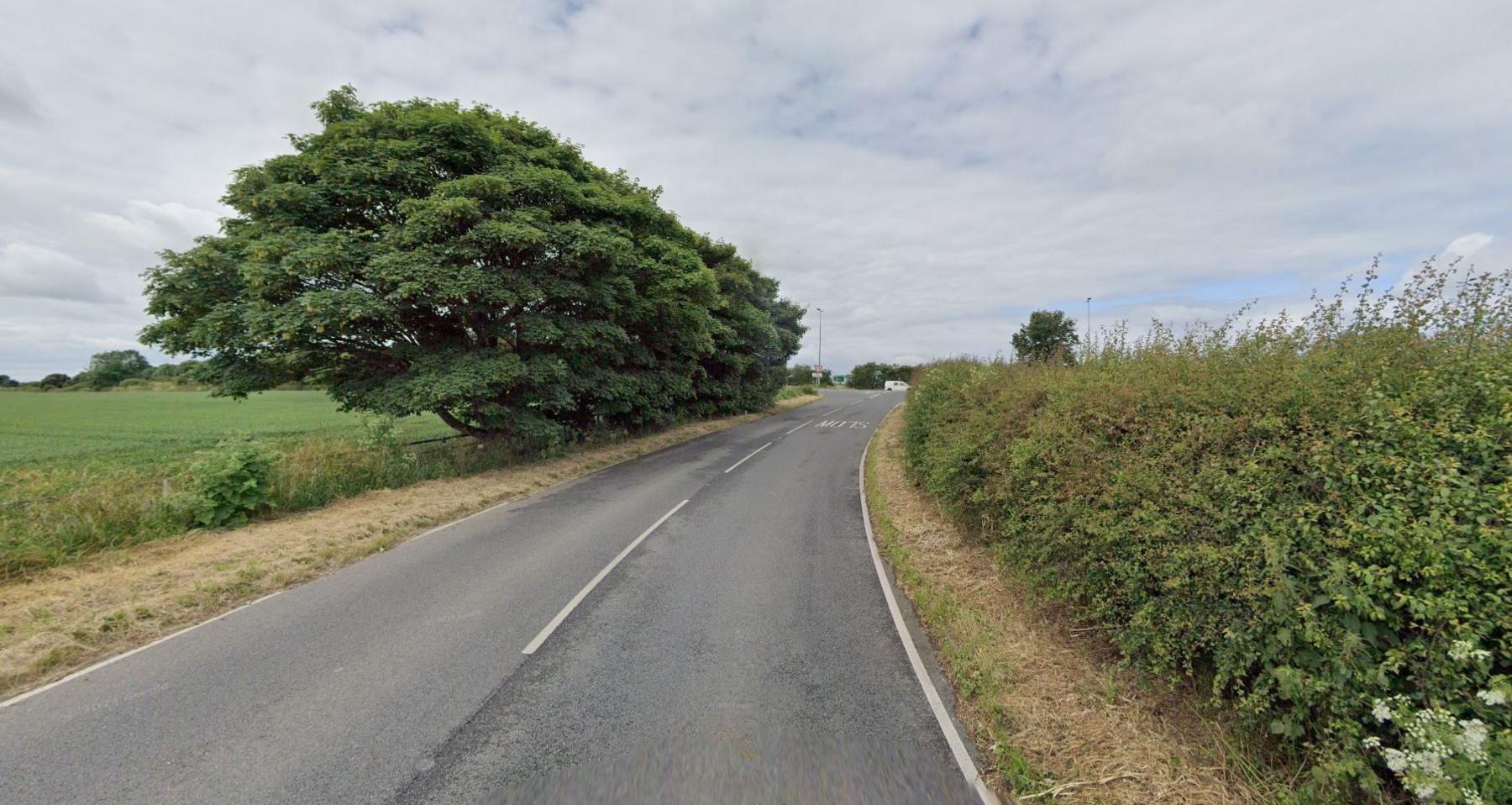 A rural single carriageway road leading to a A road junction. Trees, fields and hedges border the road on each side. The word: 'Slow' is painted in white on the right hand side.