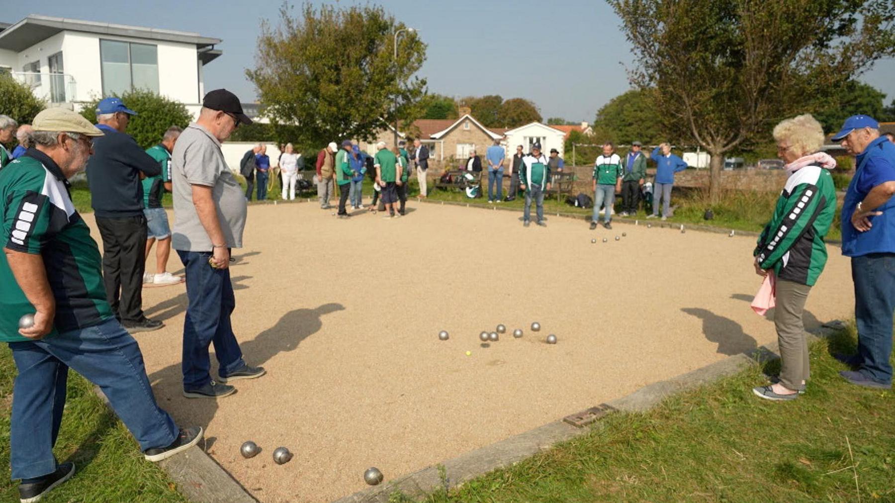 A large number of people stand around a petanque court while playing several games of the sport with the silver-coloured balls dotted across the ground.