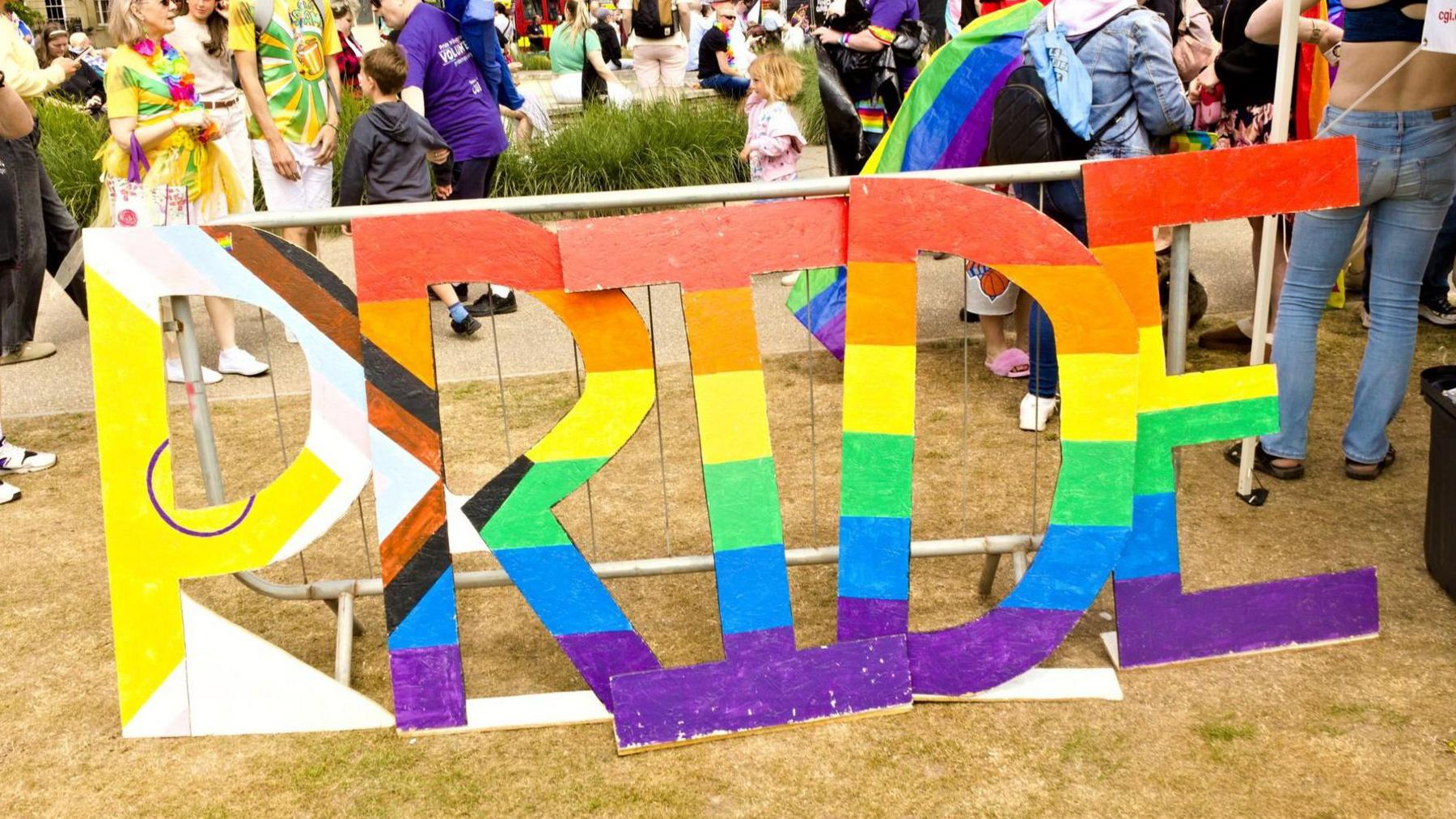 A multi-coloured sign which says "Pride" is standing on the grass with people milling behind it.
