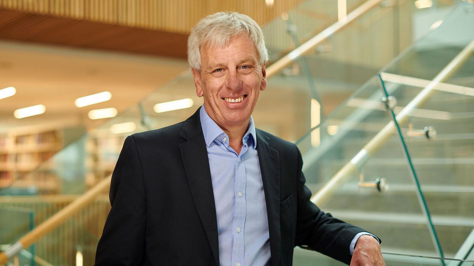 Dr John Cater smiles at the camera, wearing a black blazer and a light blue shirt, he has his left arm leaning on a stair banister as he poses for the camera in a university building. 