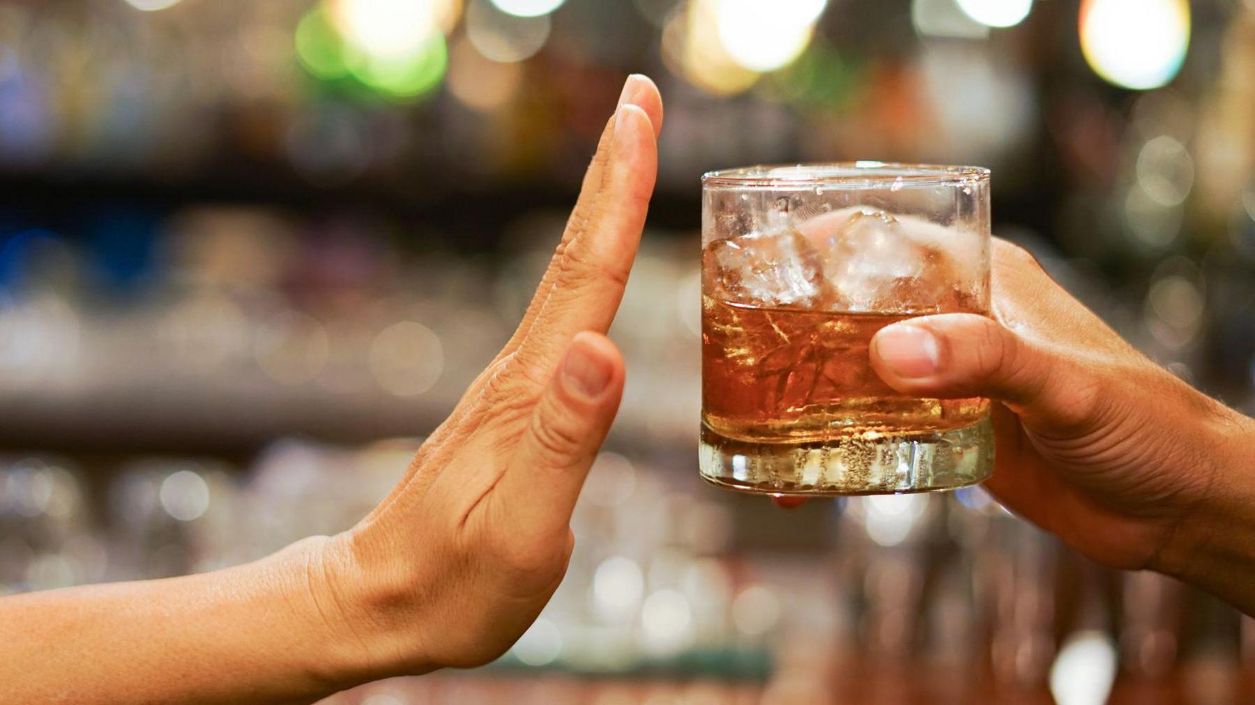 A glass of an alcoholic spirit with ice inside is offered across a bar, but a hand is outstretched as if to say stop, or no thanks.