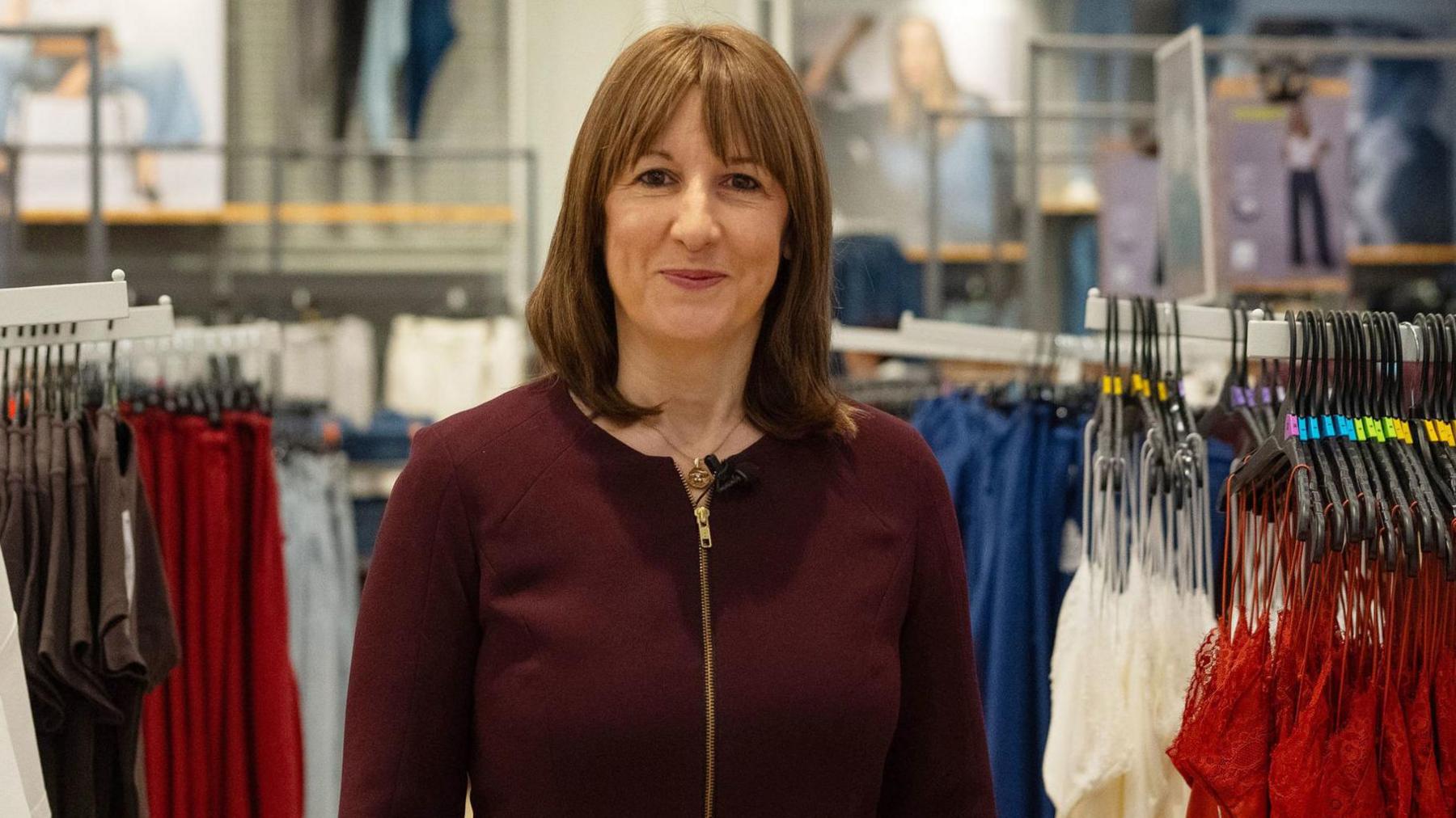 Chancellor Rachel Reeves stands in a clothes shop. She wears a burgundy jumper.