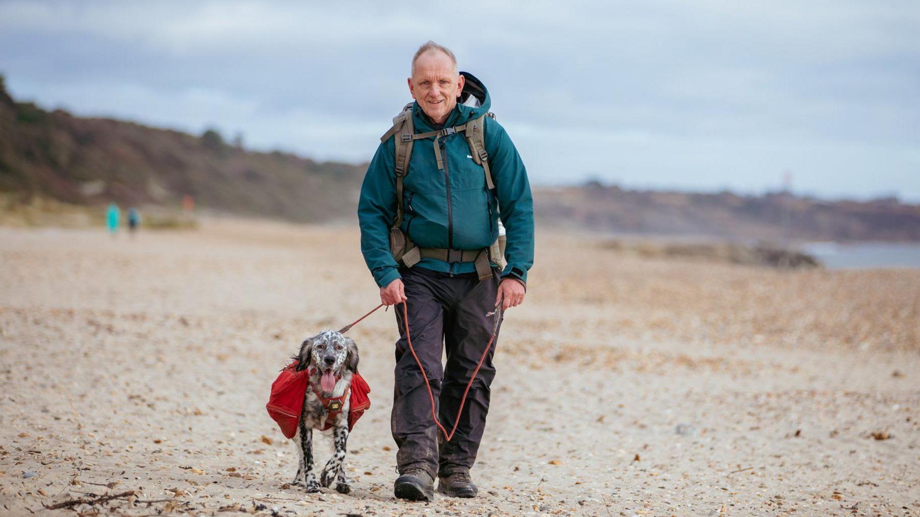 Nick and Louis walk towards the camera on a wide sandy beach, strewn with pebbles. They are both looking at the camera, and Nick is smiling while holding Louis on the lead.