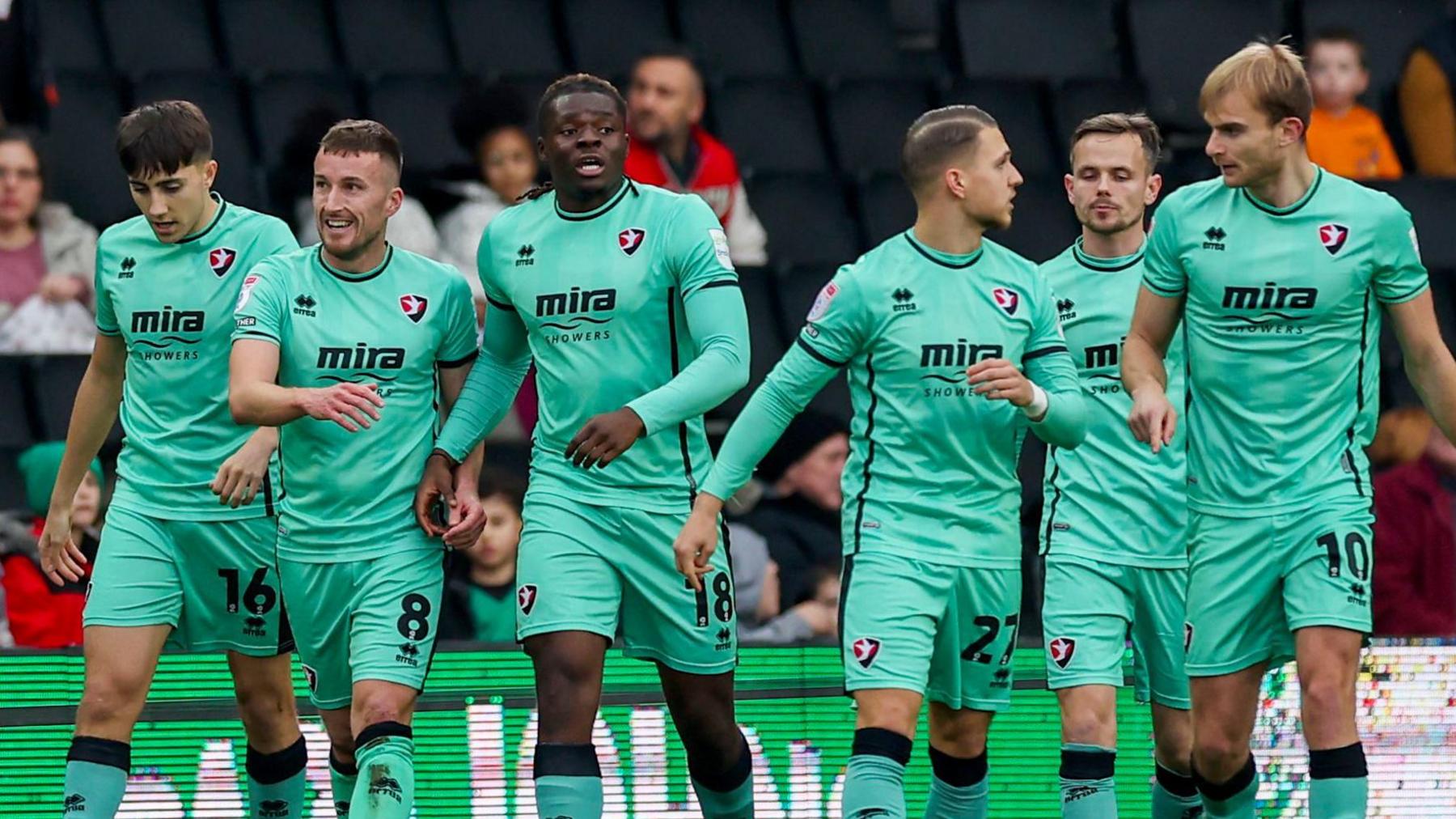 Six Cheltenham Town players on the pitch during a match against Milton Keynes Dons, wearing their turquoise away kit. They are talking to one another and smiling.