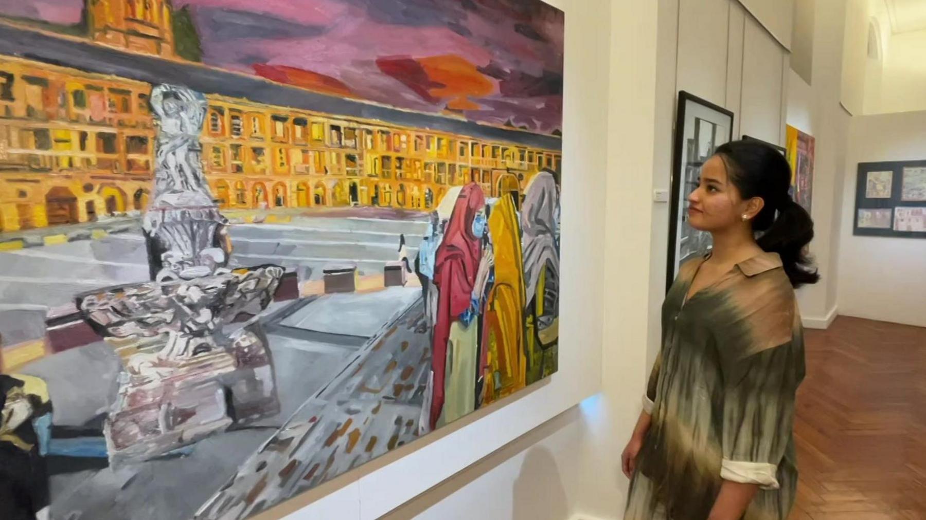 Andrea Neeta Anto stands to the right of a large painting on a wall. The painting is colourful and portrays a large Indian building with people in front of it.