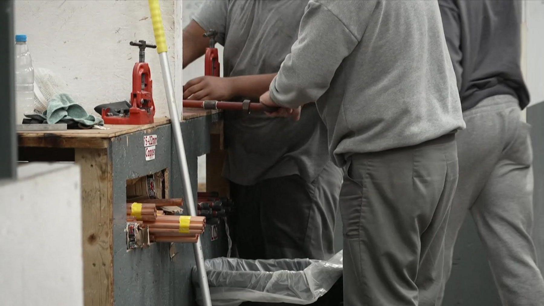 Three anonymous male prisoners wearing grey clothing operate trade tools.