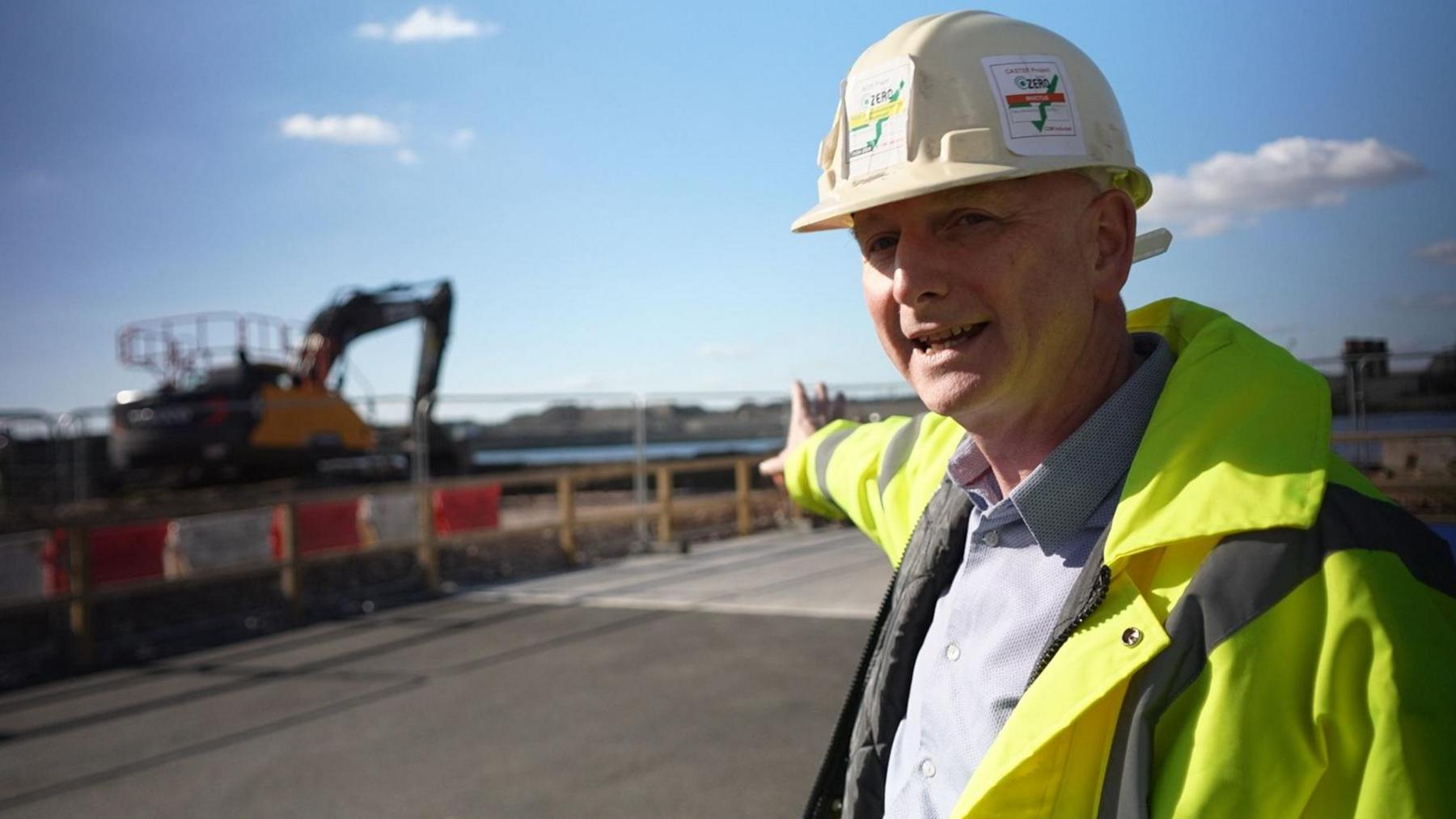 A man in a white hard hat and high-visibility jacket stands on a construction site, pointing toward an excavator in the background. Safety barriers line the area, and the sky is partly cloudy.