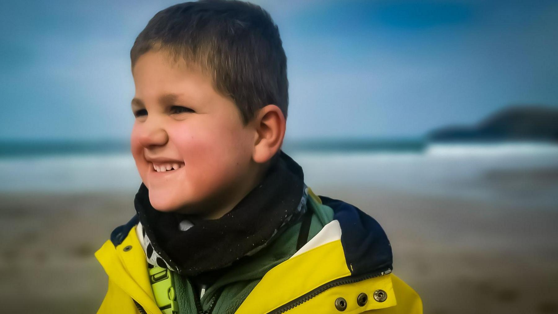Ivor looks at an angle smiling. He has short light brown hair and wearing a yellow fisherman's jacket. He is standing on beach with surf and a point in the distance